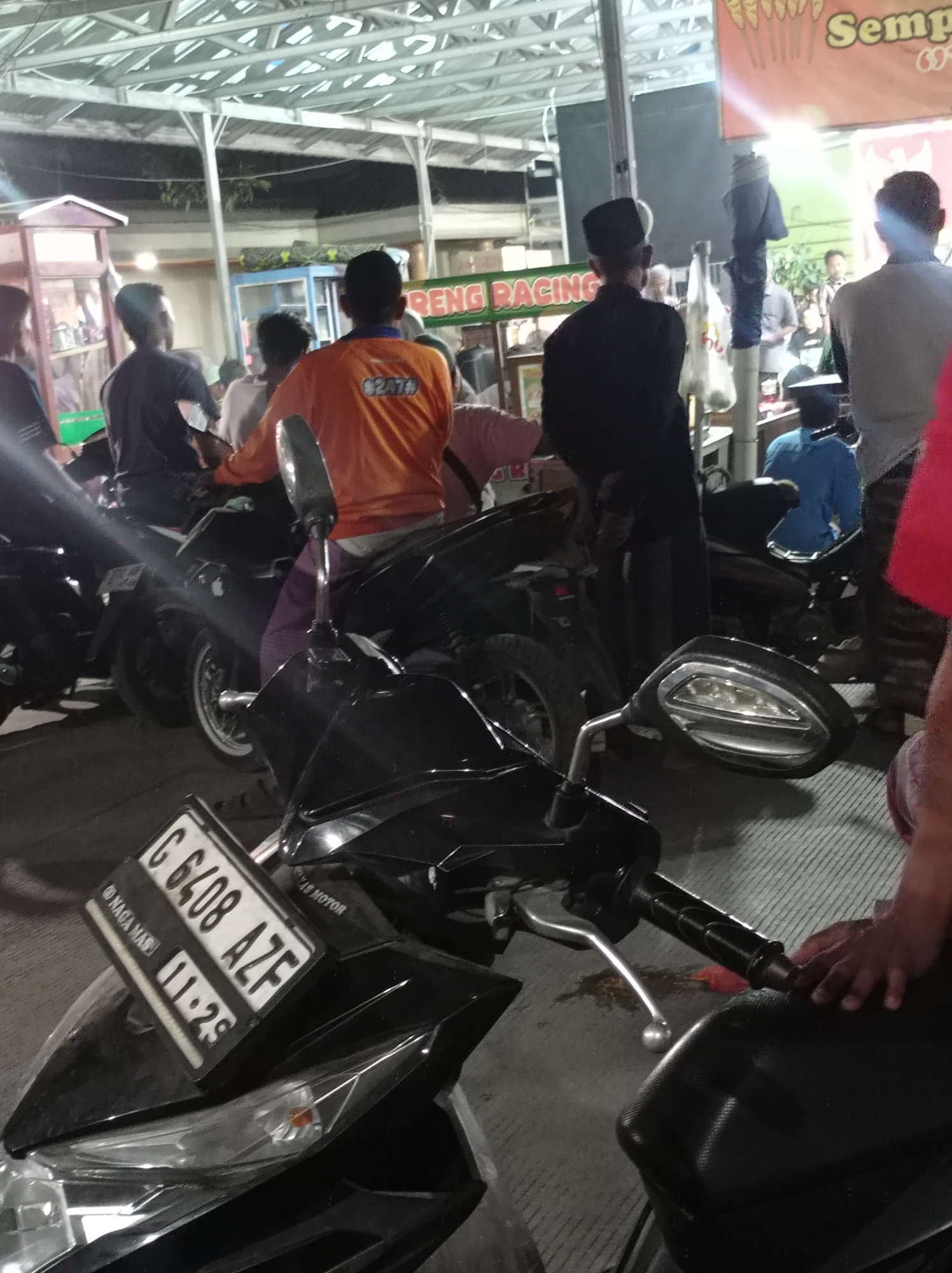 The image depicts a street scene at night, likely in Plumbungan, Indonesia, with several individuals and motorcycles. In the foreground, a black motorcycle with a white license plate reading "C 6408 AZF" and "11-29" is prominently displayed. The license plate also has the text "NAGA MAS" and "8274" visible. Behind this motorcycle, another person in an orange shirt with the number "8274" on it is seated on a motorcycle. A person in a black robe and cap stands to the right, facing away from the camera, with another person in a blue shirt seated on a motorcycle behind them. Other individuals and motorcycles are present in the background, under a metal awning. The environment appears to be a marketplace or roadside area, with some stalls or food vendors visible. The lighting suggests artificial illumination, likely from the stalls and streetlights. The ground is paved.