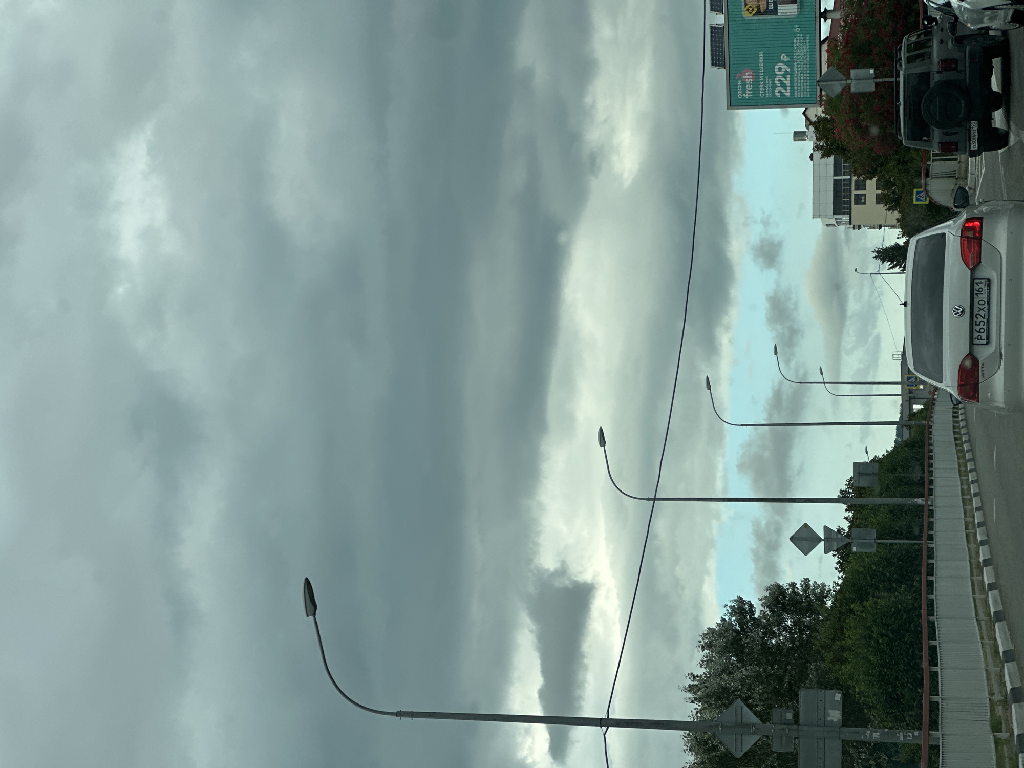 The image depicts a cloudy day in Sochi, Russia, as seen from a vehicle driving on a road. The sky is filled with heavy, gray clouds, obscuring much of the sky. Several streetlights line the road. On the right side of the road, a billboard with green and white text advertises "fresh" and shows a price of "229 ₽". Various vehicles, including a white Volkswagen sedan and a dark-colored SUV, are visible on the road. Lush green trees and bushes flank the road, with some buildings visible in the background. The overall impression is that of a typical urban scene under overcast weather conditions.