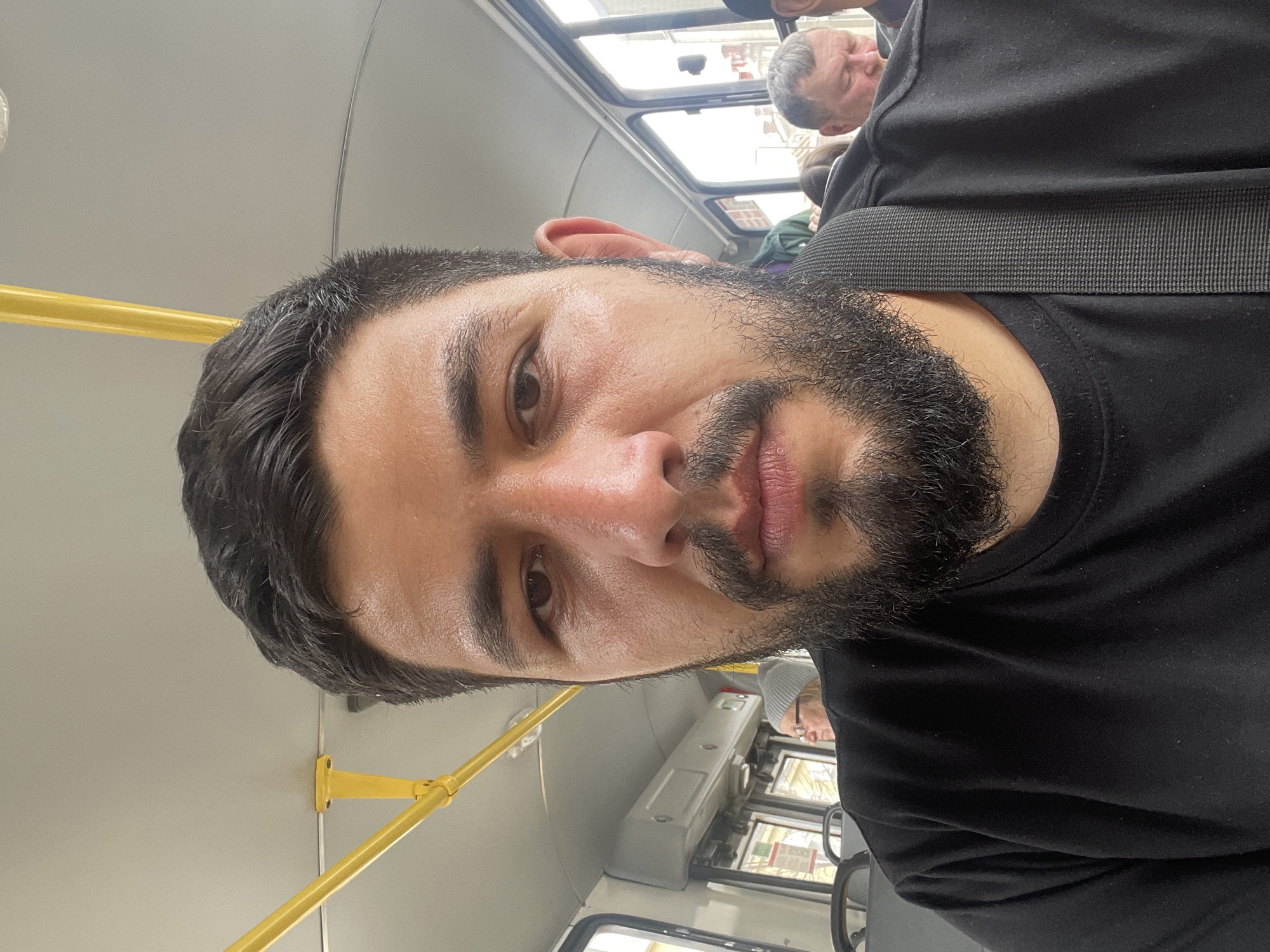 A selfie-style portrait shows a man inside a public transport vehicle, likely a bus or trolleybus, in Krasnoyarsk, Russia. The man, the primary subject, has dark hair, a full dark beard and mustache, and a medium complexion that appears slightly oily or sweaty. He is wearing a plain black t-shirt and a black shoulder strap, possibly from a bag. He looks directly at the camera with a neutral expression.
The background reveals the interior of the vehicle, characterized by light grey walls and ceiling, and bright yellow metal handrails extending horizontally across the left side of the frame. Through the windows in the upper right, daylight and elements of the outside, such as buildings and sky, are visible, indicating it is daytime.
Other passengers are partially visible in the background. An older man with short, grey hair and a neutral expression can be seen in the upper right. Further back, another person's head is partially obscured. In the lower right, part of a driver's console or dashboard is visible, showing control panels and possibly indistinct maps or diagrams, though no text is clearly legible. The scene depicts a routine urban commute.
