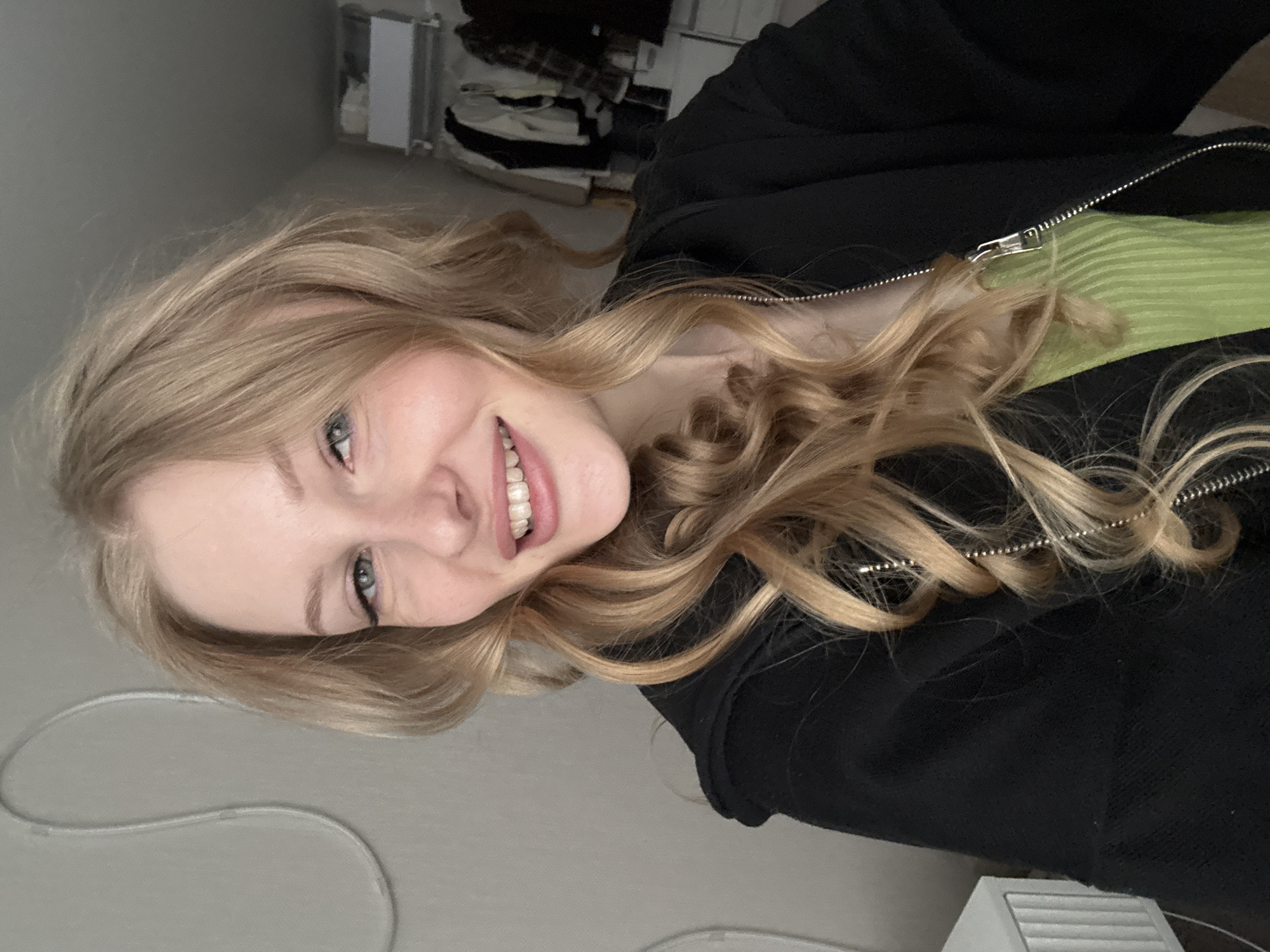 A young Caucasian woman with blonde wavy hair, blue eyes, and fair skin is shown from the chest up. She is wearing a black zip-up hoodie over a green ribbed top. She is smiling, revealing her teeth, and looking towards the camera. Her hair is styled with loose curls, framing her face. In the background, there is a grey wall with a white curved decoration, a dresser or cabinet on the left, and a closet with clothes on hangers on the right. The lighting appears to be natural and soft, suggesting an indoor setting. The overall scene is casual and personal. The location is identified as Perm, Russia.