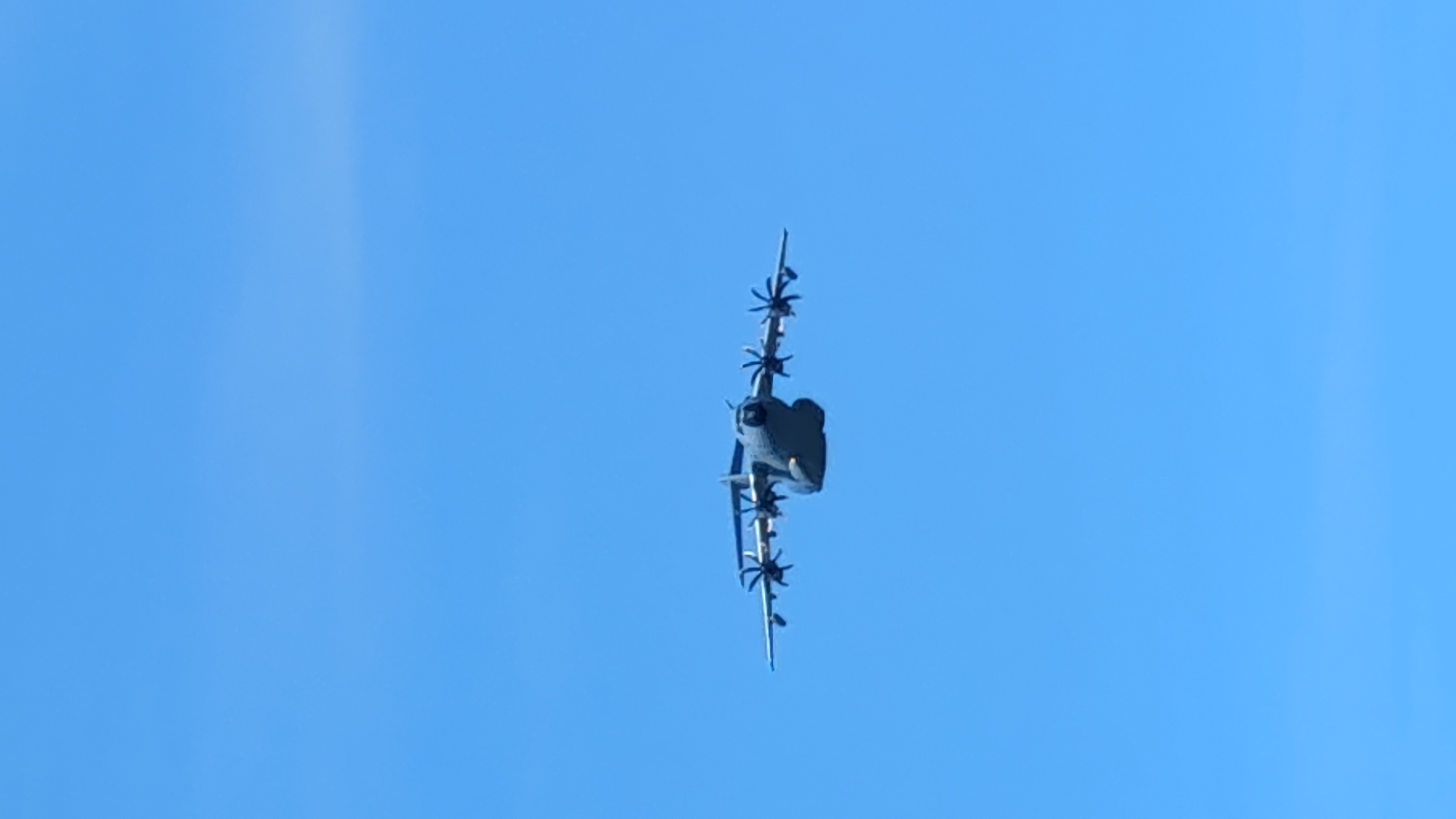 An Airbus A400M Atlas military transport aircraft is captured in flight against a clear blue sky. The aircraft is viewed from a frontal perspective, slightly from below, highlighting its four turboprop engines, distinctive high wings, and bulky fuselage. The propellers on the engines appear to be in motion, suggesting the aircraft is actively flying. The bright blue sky dominates the image, with no visible clouds or ground details. The image does not provide any specific visual cues to indicate its location in Warsaw, Poland.