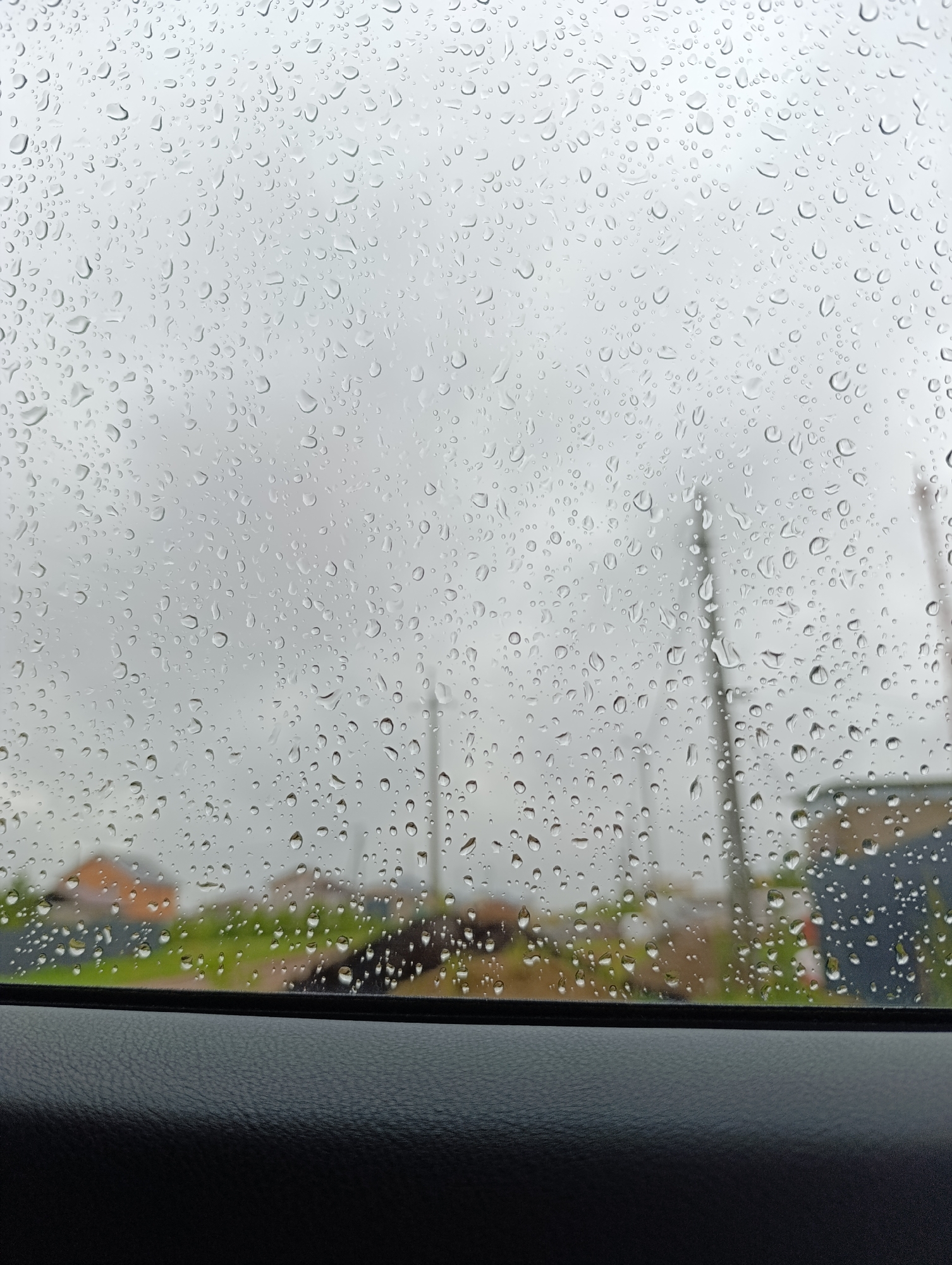 The image is a view through a rain-covered window, likely a car window given the black dashboard-like surface in the foreground. Raindrops obscure the background, which features indistinct structures that may be houses or buildings. There are also utility poles visible in the background. The weather appears overcast and gloomy. Considering the location, the background structures and utility poles are suggestive of a residential or suburban area on the outskirts of Chaikovsky, Russia.