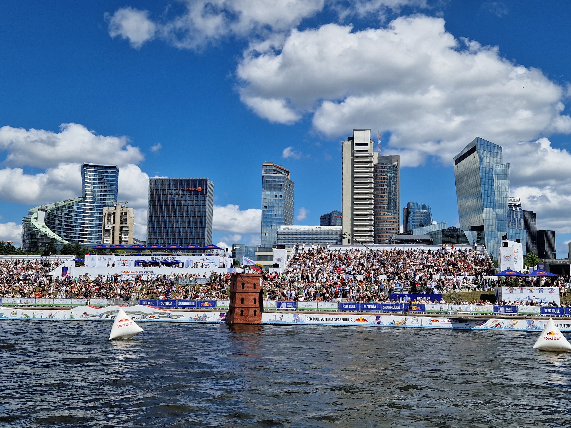 The image depicts a Red Bull event taking place on the Neris River in Vilnius, Lithuania. The scene is set against the backdrop of the modern Vilnius cityscape, featuring several contemporary skyscrapers, including the Swedbank building. In the foreground, the dark water of the river is visible, with a white triangular buoy bearing the Red Bull logo floating on the surface. Behind the river, a large grandstand filled with spectators is set up. The grandstand is adorned with various Red Bull branding and banners, and at the front edge, a decorative brick tower stands in the water. The sky is a vibrant blue with scattered white clouds.