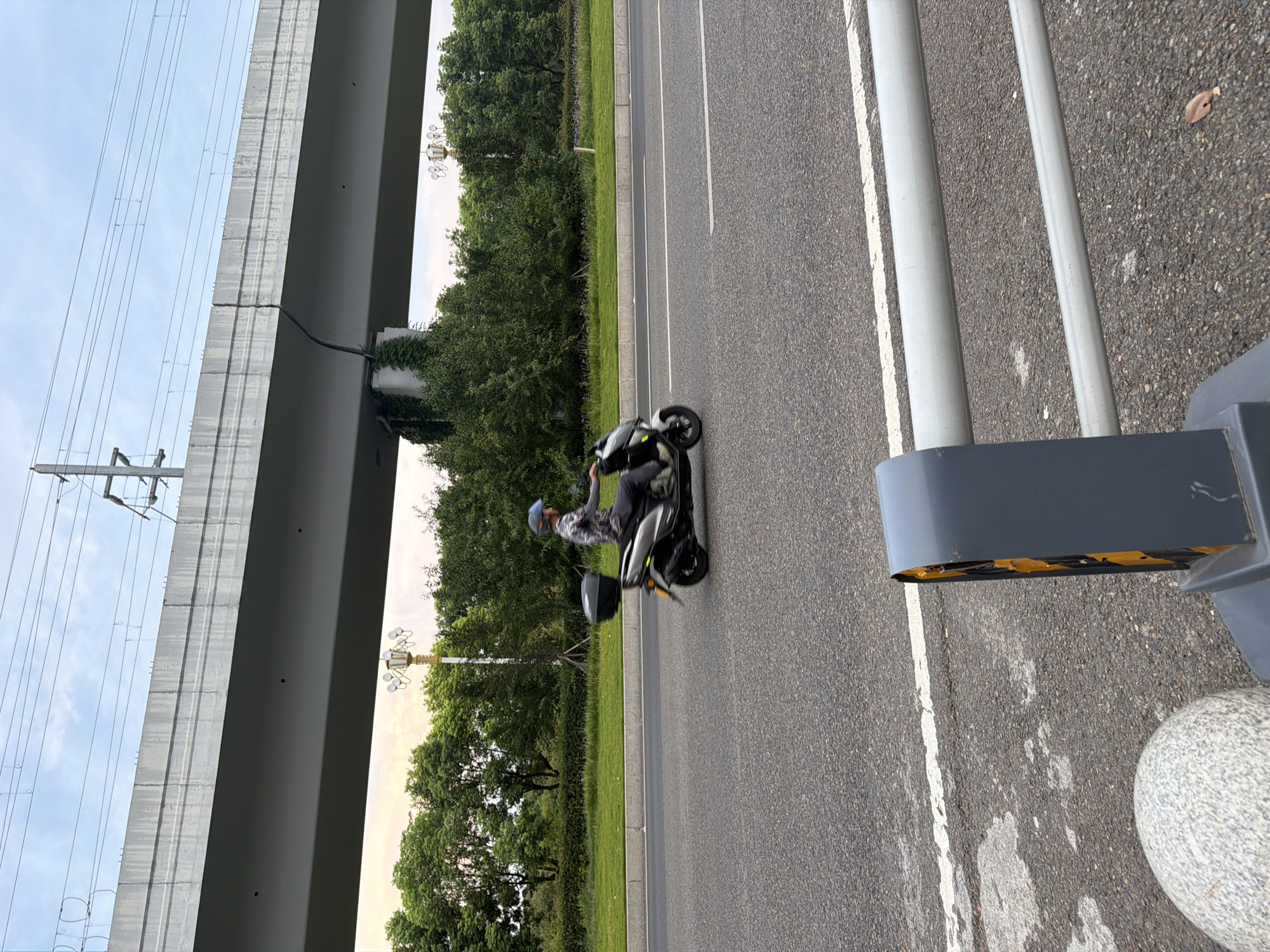 A person rides a scooter on a road under an elevated structure. The scooter is silver and black, with a black storage box on the back. The rider is wearing a camouflage shirt, dark pants, and a blue helmet. The road is asphalt, with white lines marking lanes. There are trees and grass on the side of the road, and a large concrete structure above. The sky is cloudy. In the foreground, there is a grey barrier with yellow accents and a round grey object. The scene appears to be in a city environment with infrastructure elements like a road and an overpass.