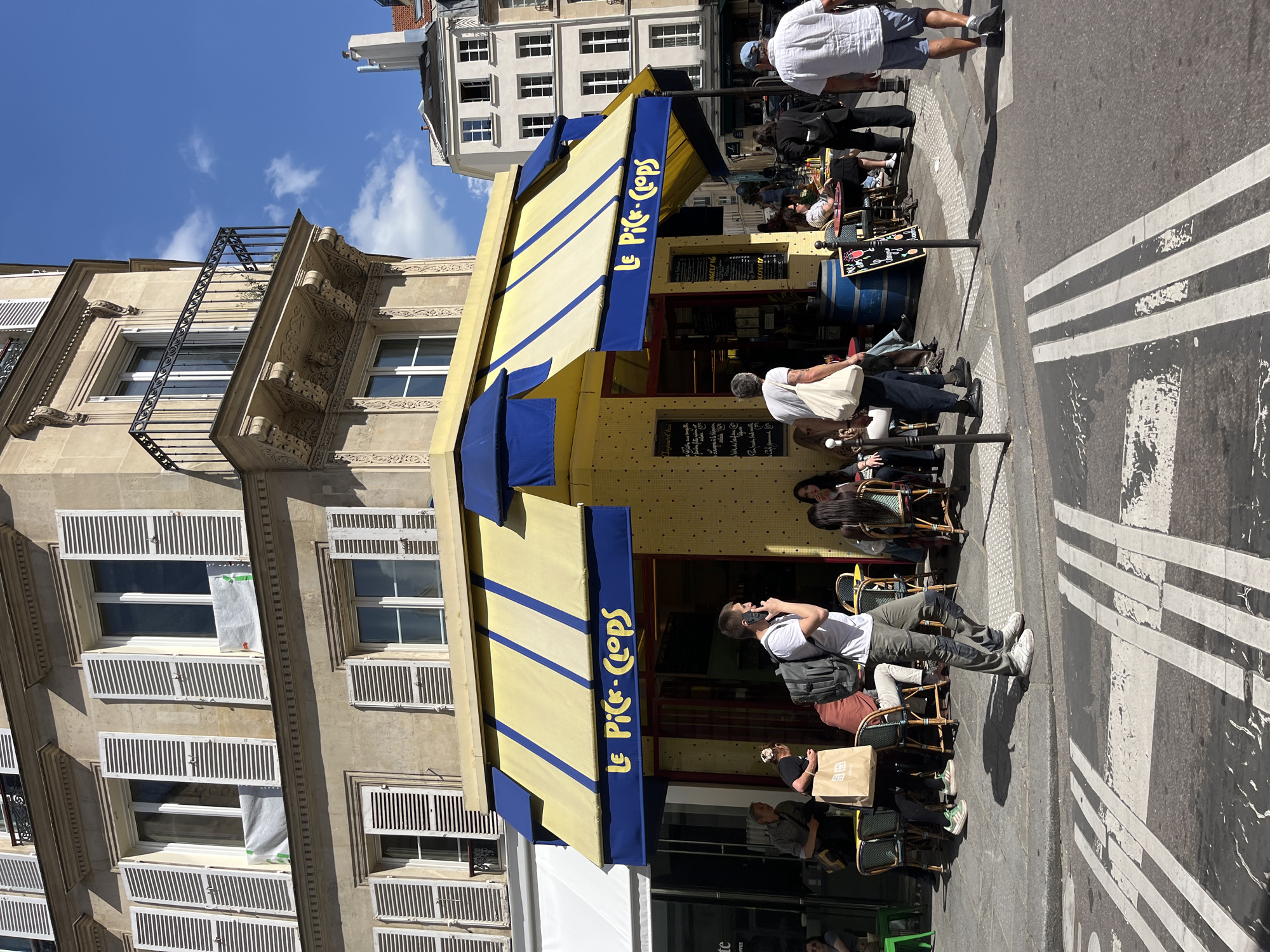 The image depicts a street scene in Paris, France. The primary subject is "Le Pic-Clops," a restaurant or cafe, identified by its yellow awning with blue trim, prominently displaying the name in blue lettering. Several people are seated at tables outside the establishment, suggesting active patronage. A man stands to the side, appearing to be using a mobile phone. Pedestrians are also seen walking on the sidewalk. The restaurant is situated on a street marked with white pedestrian crossing stripes. The architecture of the building housing the cafe is characterized by light-colored stone, ornate detailing, and shuttered windows. The sky is blue with scattered clouds, indicating a sunny day.