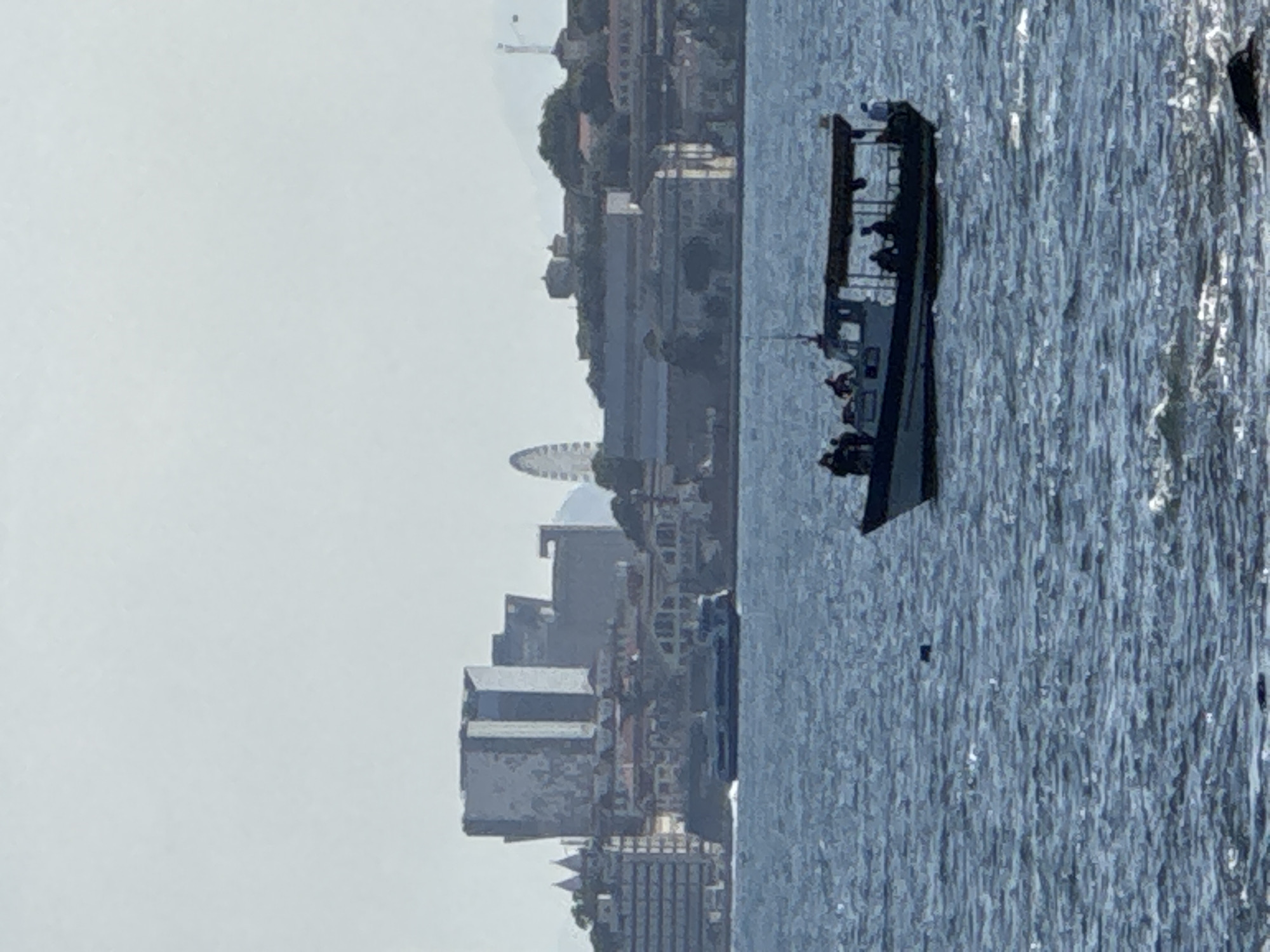 The image captures a wide shot of a body of water with a cityscape in the background. In the foreground, a motorboat carrying several passengers is navigating the rippling blue water. The boat has a covered seating area and appears to be moving to the right. In the mid-ground, a larger ferry or passenger boat is seen moving left in front of the city's waterfront.
The city skyline features a variety of buildings, including modern high-rises and older structures. A Ferris wheel is visible in the distance on a hill, suggesting an amusement area or landmark. The architecture suggests a port city with a mix of contemporary and older development. The overall scene is set against a hazy, light-colored sky, indicating a bright but possibly overcast day. The image is taken from a low vantage point, looking across the water towards the city. This scene is likely of Niterói, Brazil, given the prominent architecture and the waterfront setting which is characteristic of the city's coastline along Guanabara Bay.