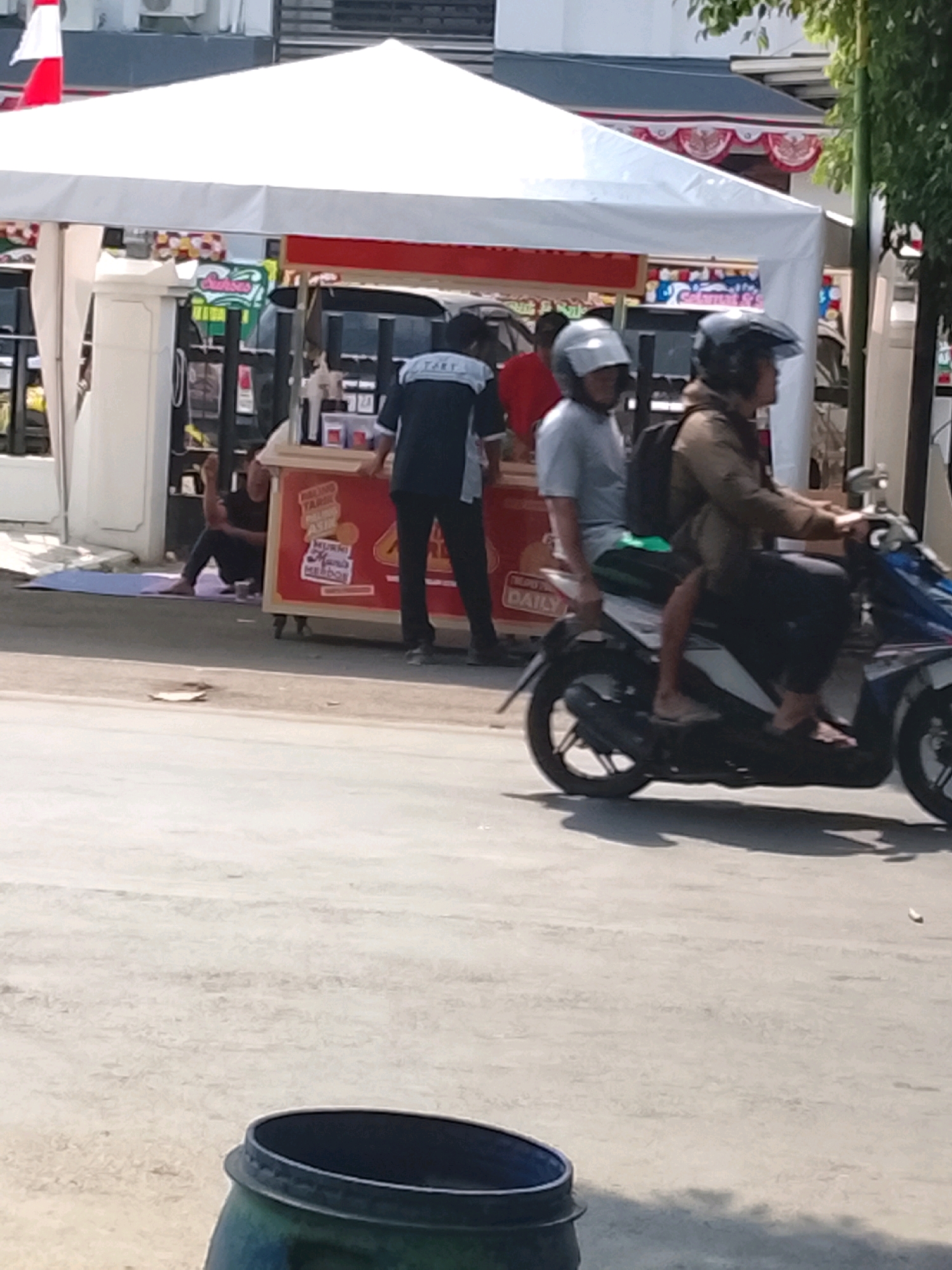 In the city of Slawi, Indonesia, a street scene unfolds under a white tent, likely a temporary stall or market. Two men are riding on a scooter. The driver, wearing a brown jacket and black pants, has a backpack. The passenger, in a grey shirt and black pants, wears a white helmet. In the background, a vendor in a dark blue uniform with a "TART" logo on the back is seen. Another person is sitting on a purple mat on the ground. The stall displays red and yellow signage, with some text visible including "DAILY." The environment is an outdoor setting with a paved ground. In the foreground, a dark colored barrel is partially visible.