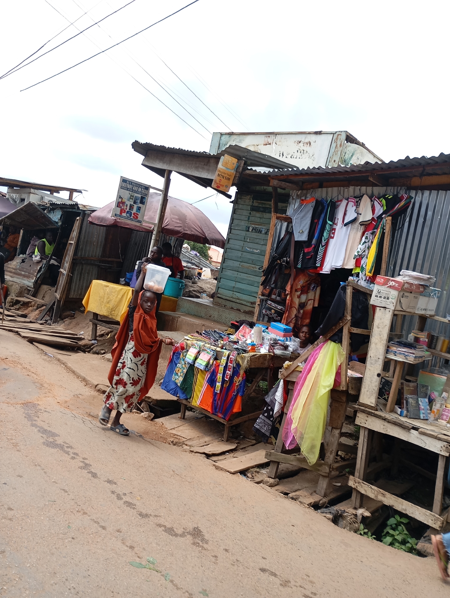 In Jos, Nigeria, a bustling market scene unfolds on a dusty street. A young girl, wearing a patterned dress and an orange head covering, walks along the road, carrying a clear plastic container filled with eggs on her head. She is moving past a makeshift stall displaying an array of colorful clothing items, including t-shirts and shorts, neatly hung on a rack. The stall itself is constructed from corrugated metal and wood, with various goods piled on shelves and tables. Further down the street, other stalls and structures are visible, suggesting a vibrant commercial area. The sky above is overcast, hinting at a potentially humid or rainy day. The overall impression is one of everyday life and commerce in a Nigerian city.