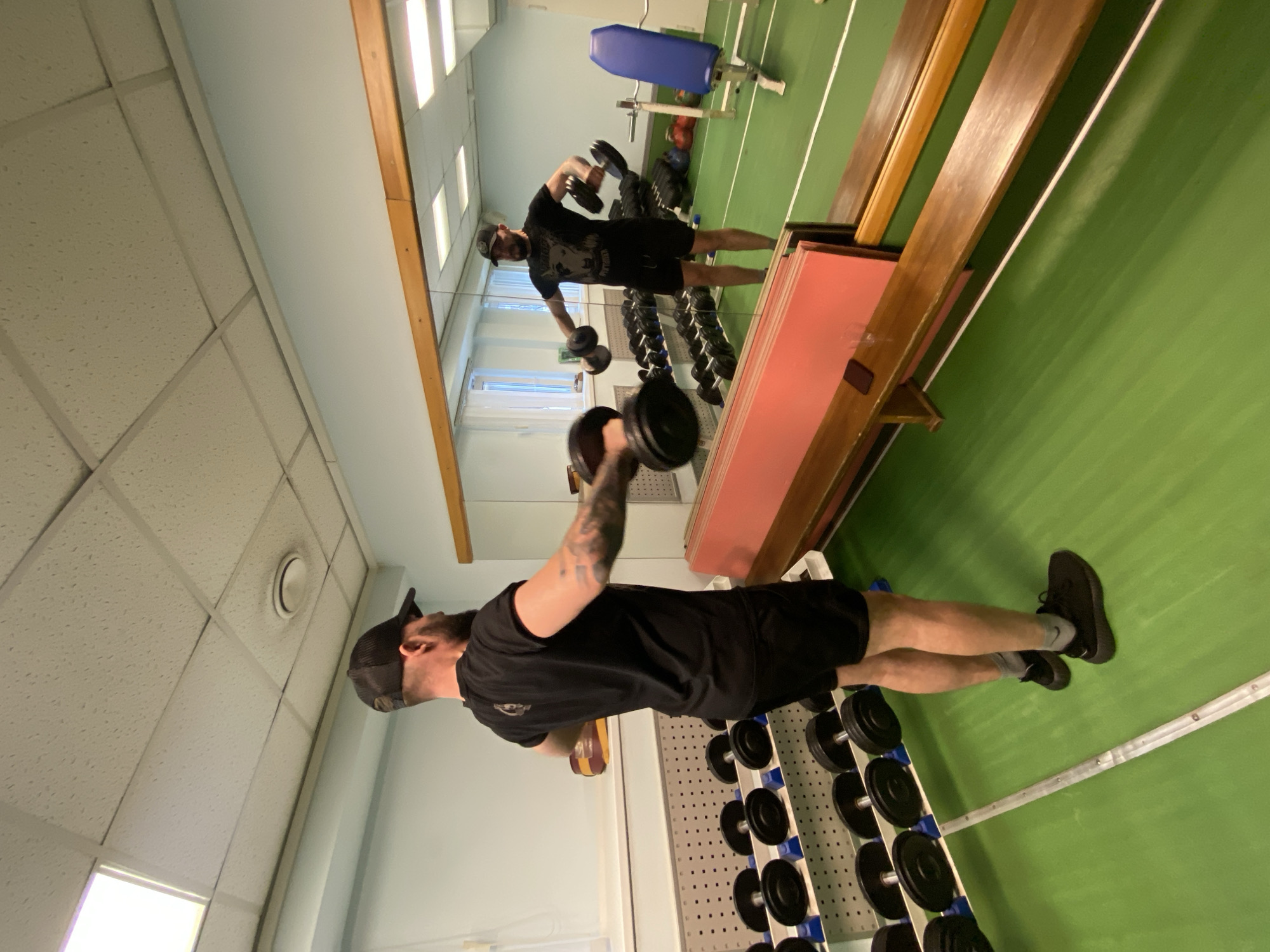 A man is performing a lateral dumbbell raise in a gym in Chaikovsky, Russia. He is wearing a black T-shirt, black shorts, a baseball cap, and black sneakers. He is standing near a rack of dumbbells and facing a mirror. The gym has a green floor, a white ceiling with fluorescent lights, and blue exercise equipment. His reflection can be seen in the mirror, suggesting he is using the mirror to monitor his form. The mirror is framed by wooden trim.
