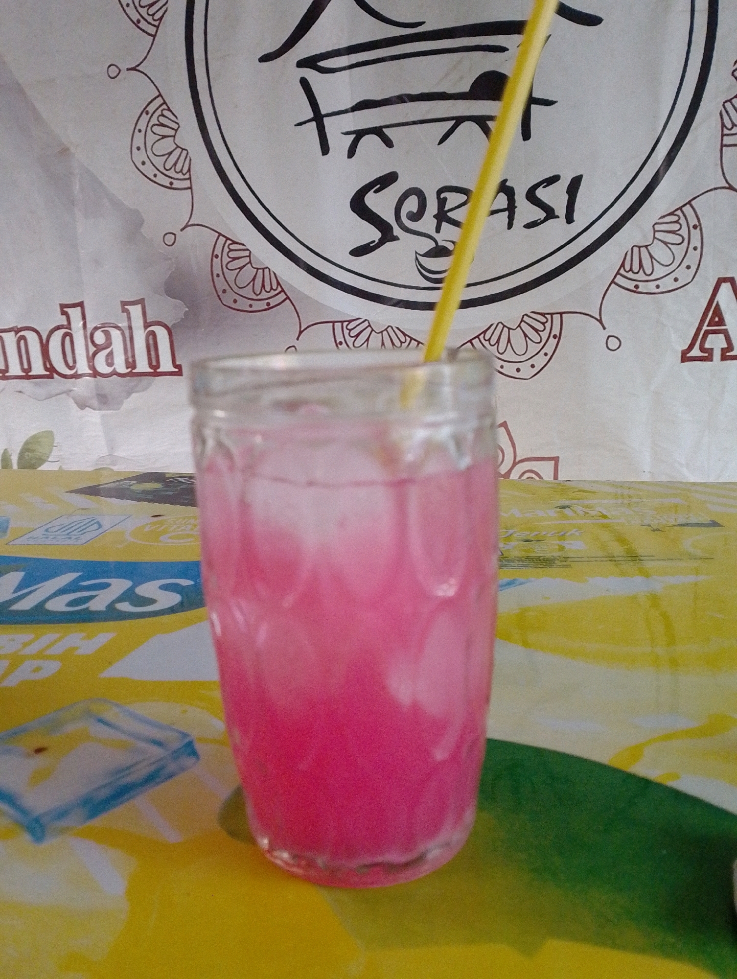 A close-up, eye-level, vertical shot captures a glass filled with a pink beverage and ice, adorned with a yellow straw. The glass sits on a table covered with a brightly colored, patterned tablecloth featuring yellow, blue, and green accents. In the background, a banner displays the text "SORASI" in a stylized font, framed by decorative elements. The banner also shows partial text such as "indah" and the letter "A". This scene is set in Ngeling, Indonesia.