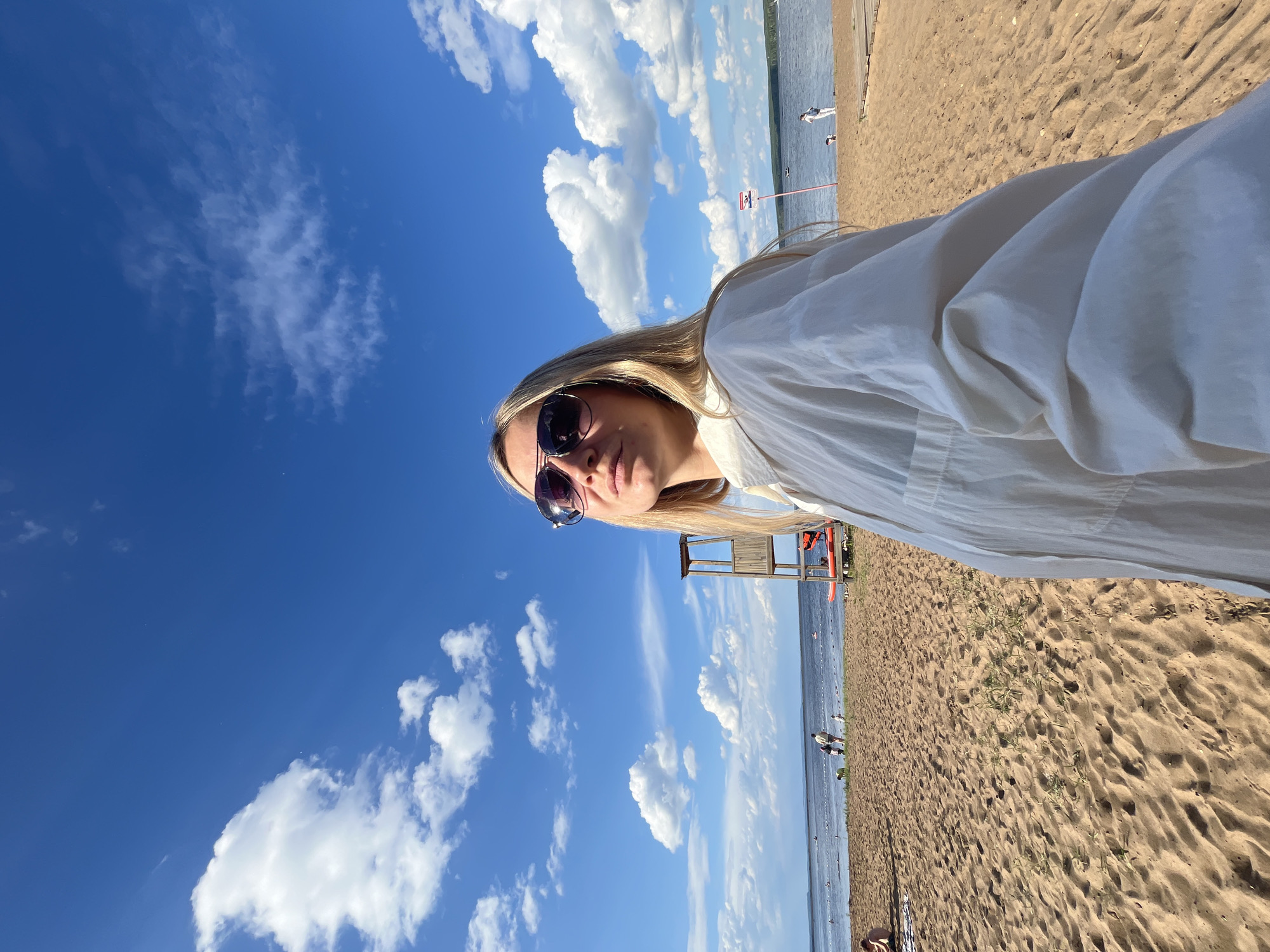 The media file depicts a woman taking a selfie on a sandy beach. The woman has blonde hair and is wearing sunglasses and a white shirt. She is looking at the camera with a relaxed expression. The background shows a bright blue sky filled with fluffy white clouds. To the right of the woman, there is a lifeguard tower and a body of water, likely a lake or river, with a few people visible in the distance. The sand is tan and textured, with some sparse green vegetation growing on it. The overall setting suggests a sunny day at the beach. The image is oriented vertically.
