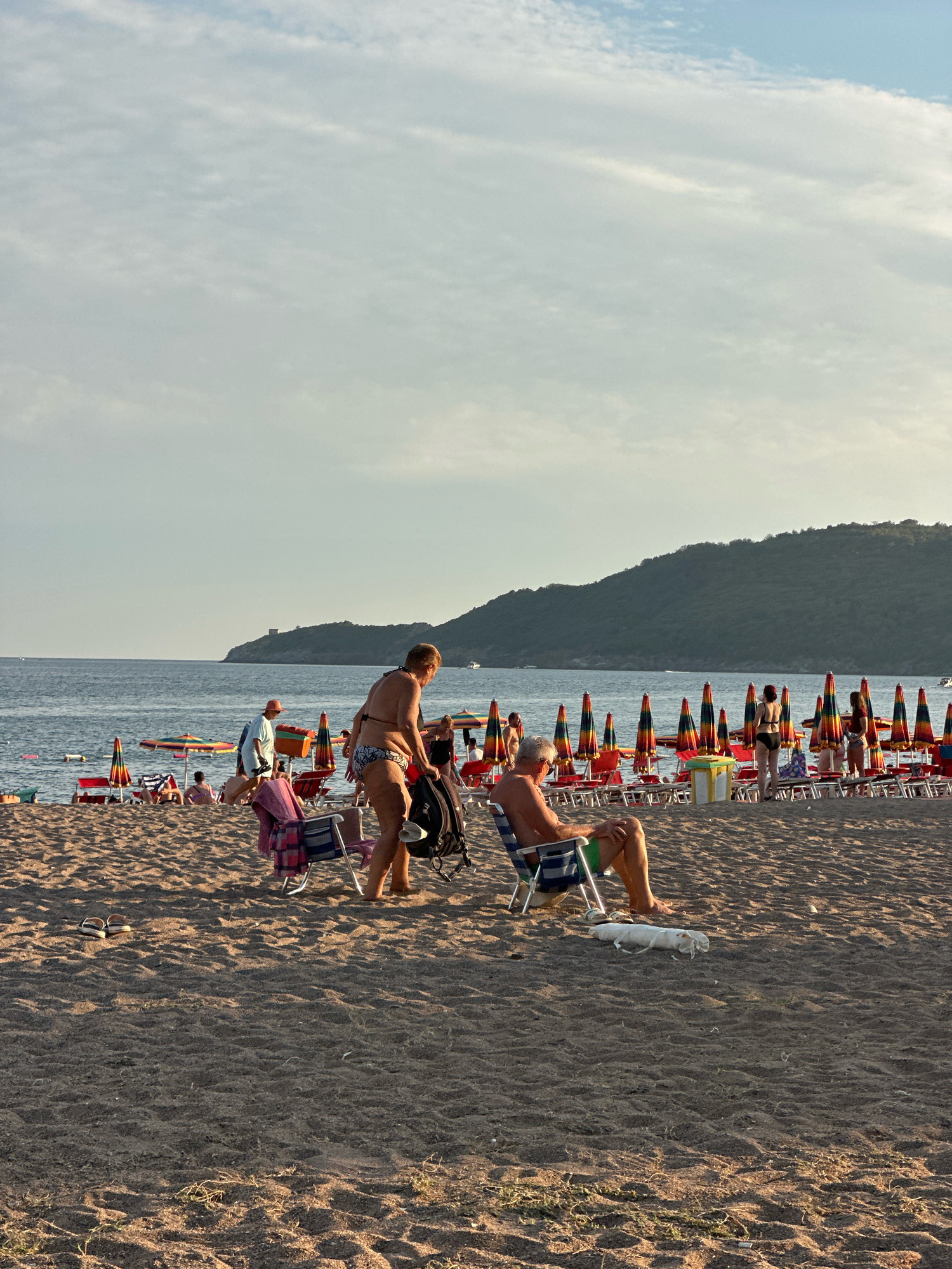 The image shows a crowded beach scene on a sunny day, likely in or near Boreti, Montenegro, given the hilly coastline in the background. The foreground is dominated by a sandy beach with rows of colorful umbrellas and beach chairs. Several people are visible, including a man in a blue and white striped chair on the right, relaxing with his legs extended. A small white dog lies on the sand in front of him. To the left, a woman in a bikini is standing and appears to be adjusting a backpack. Further back, other beachgoers are scattered among the chairs and umbrellas, with some wading in the calm sea. The water is a deep blue, meeting the horizon with a hazy sky above. The overall atmosphere is one of leisure and summer vacation.