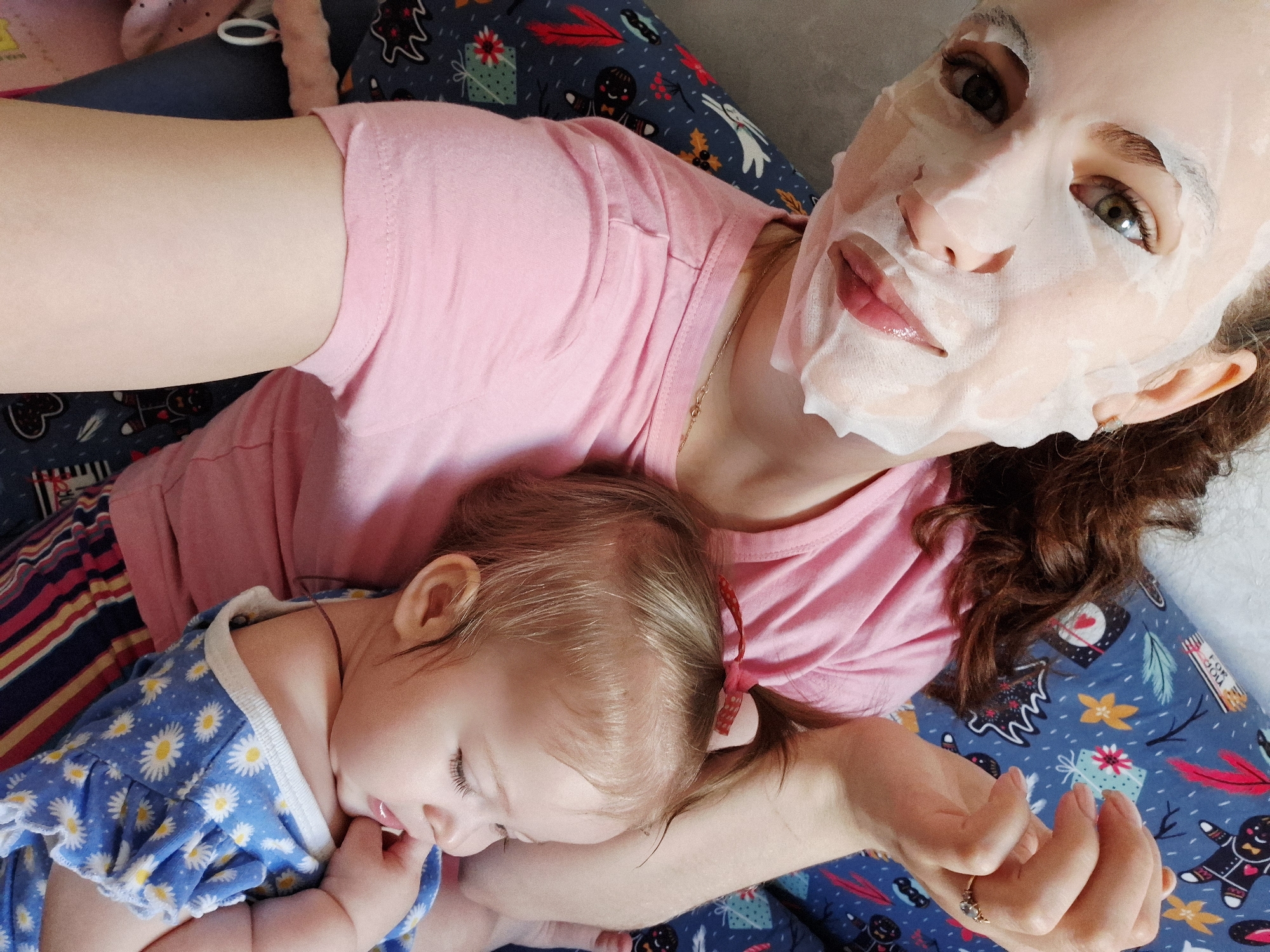 A woman wearing a pink t-shirt and a sheet mask on her face lies on a bed with festive patterned blue and white bed sheets. She cradles a sleeping baby wearing a blue and white daisy-patterned outfit. The baby has a red bow in her hair and a pacifier in her mouth. The woman is holding her hand. The location is Chaikovsky, Russia.