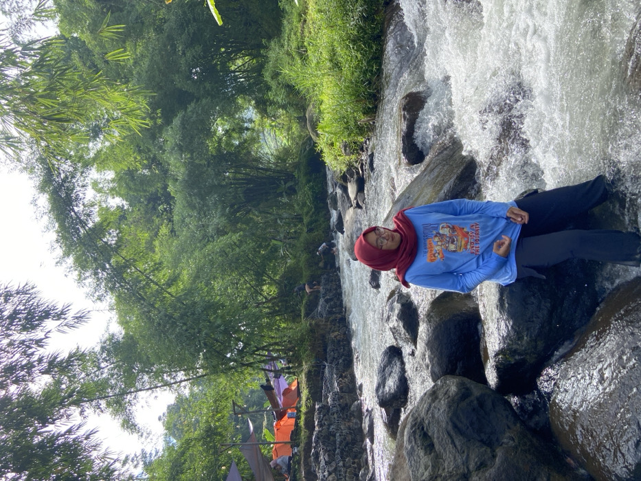 A woman in a hijab, blue long-sleeve shirt, and dark pants stands on rocks in a shallow, fast-moving stream surrounded by lush green vegetation. Tall bamboo trees are visible on the left side of the frame. Tents can be seen pitched near the stream, suggesting a campsite. The woman faces the viewer, partially immersed in the water. The surrounding environment is predominantly green with rocky elements. The lighting is bright and natural.