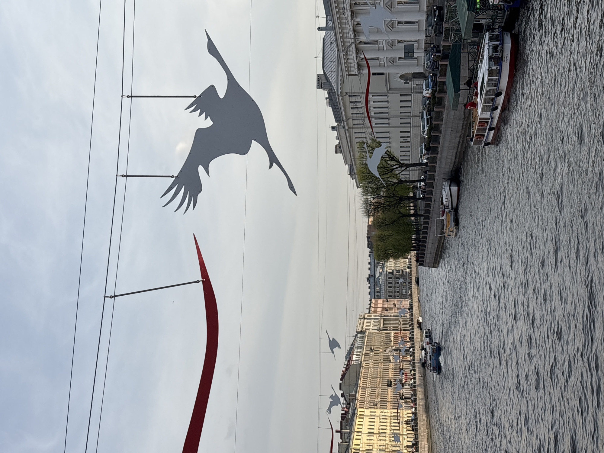 The image depicts a canal in Saint Petersburg, Russia, adorned with artistic decorations consisting of stylized crane silhouettes and red accents suspended above the water. A tour boat is docked along the canal bank. Buildings line the canal, some reflecting in the water. Cars are parked along the embankment. The sky is overcast.