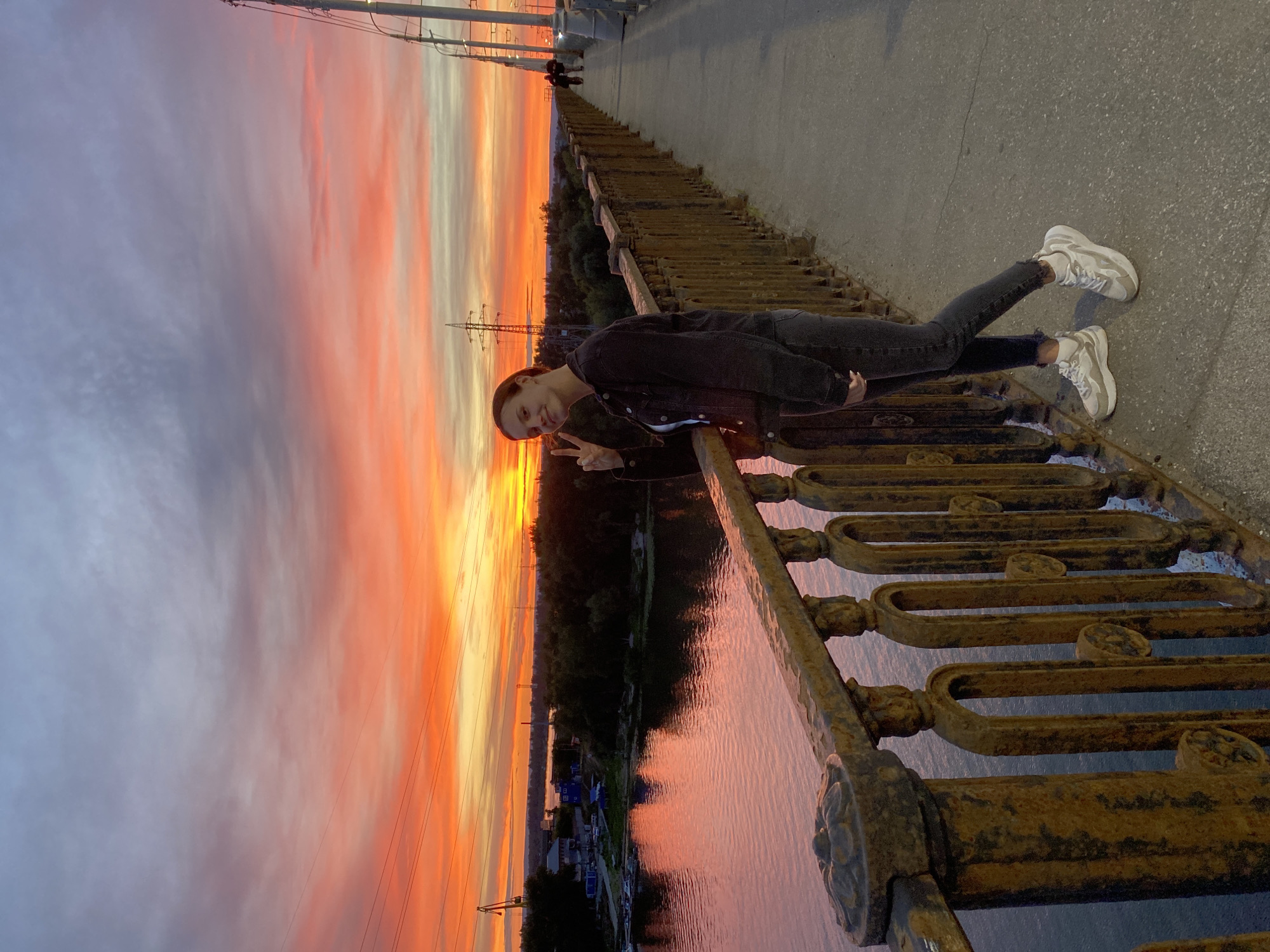 The image shows a young woman on a bridge, leaning over a rusty railing. She is wearing a dark jacket, dark pants, and white sneakers. She is smiling and making a peace sign with her fingers. The bridge has a weathered, industrial look with ornate, rusted metalwork.
Behind the woman, the sky is filled with a vibrant sunset, displaying a gradient of colors from deep orange and red to soft pink and purple, with streaks of white clouds. The sun is low on the horizon, casting a warm glow. Below the bridge, a calm body of water reflects the colors of the sky. Trees line the opposite bank of the water, with a few small buildings and what appear to be boats docked near the shore. In the distance, a large power line tower is visible against the colorful sky. The overall mood is peaceful and picturesque, with the woman enjoying the beautiful sunset. The time of day is clearly evening, indicated by the sunset. No text is visible in the image.