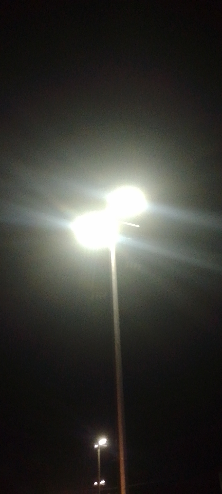 The image depicts a nighttime outdoor scene in the city of Unknown, Unknown. The primary subject is a tall, slender light pole positioned centrally in the upper half of the frame. This pole supports a luminaire featuring two distinct, intensely bright white light sources, which are overexposed and emit significant lens flare horizontally across the dark background. A secondary, smaller light pole with at least two illuminated white light sources is visible in the lower-left quadrant, appearing further in the distance and less intensely lit. The immediate environment consists of a uniformly dark, unidentifiable background, indicating low ambient light conditions. No individuals, vehicles, or specific architectural features are discernible. The image is characterized by strong artificial illumination against a dark, possibly overcast, night sky.