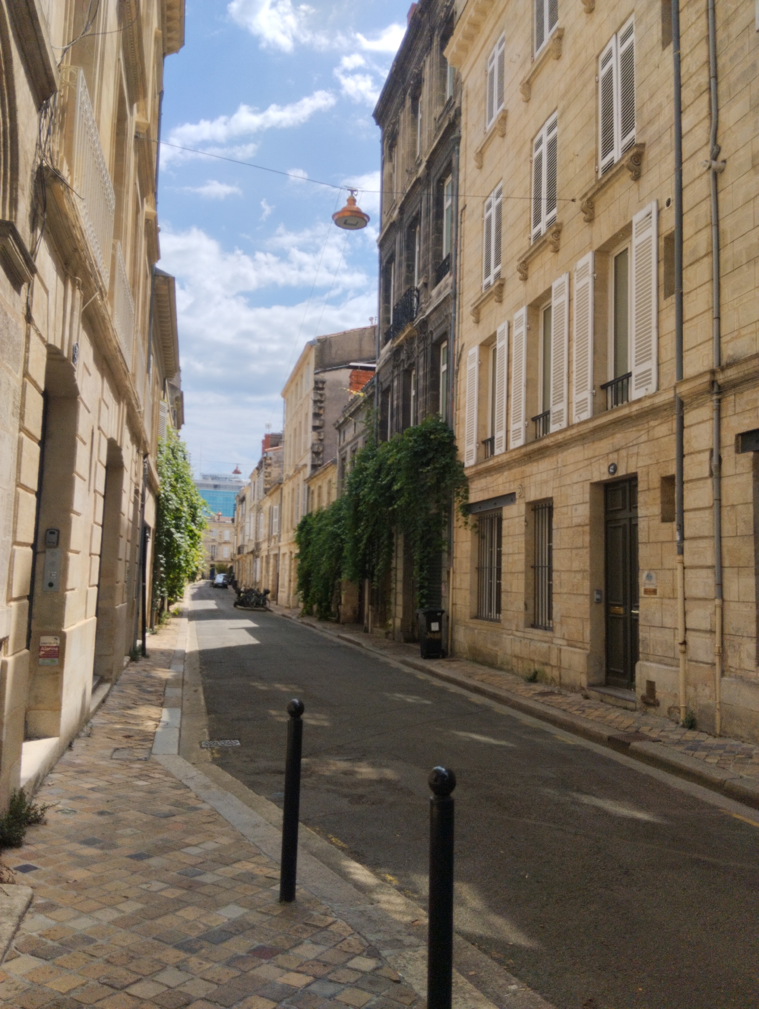 This is a street scene in Bordeaux, France, likely on a moderately cloudy day. The perspective is looking down a narrow street lined with multi-story buildings. The buildings are constructed of light-colored stone, with many windows featuring white shutters. On the left side of the street, the building is closer and its stone facade is more visible, showing details like windows and what appears to be an intercom system and an alarm sticker. The street itself is paved with asphalt, and there are sidewalks made of cobblestones. A few black bollards are positioned along the sidewalk. Vegetation, in the form of ivy or climbing plants, grows along the sides of some buildings. In the distance, the street curves slightly, and several motorcycles are parked. A modern glass-fronted building is visible beyond the older architecture. An orange street lamp hangs on a wire suspended across the street.