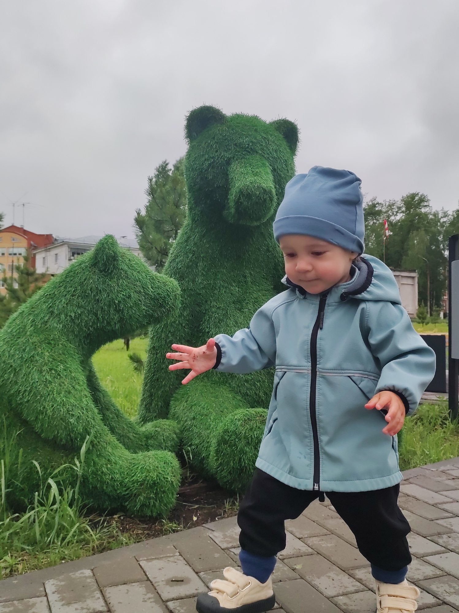 In the city of Chaikovsky, Russia, a young child stands on a brick path in front of two topiary bear sculptures. The child, wearing a blue hat and jacket over dark pants and light-colored shoes, has their hand outstretched toward the bears. The bears are made of green foliage and are positioned on a grassy area. The background includes buildings, trees, and a cloudy sky.