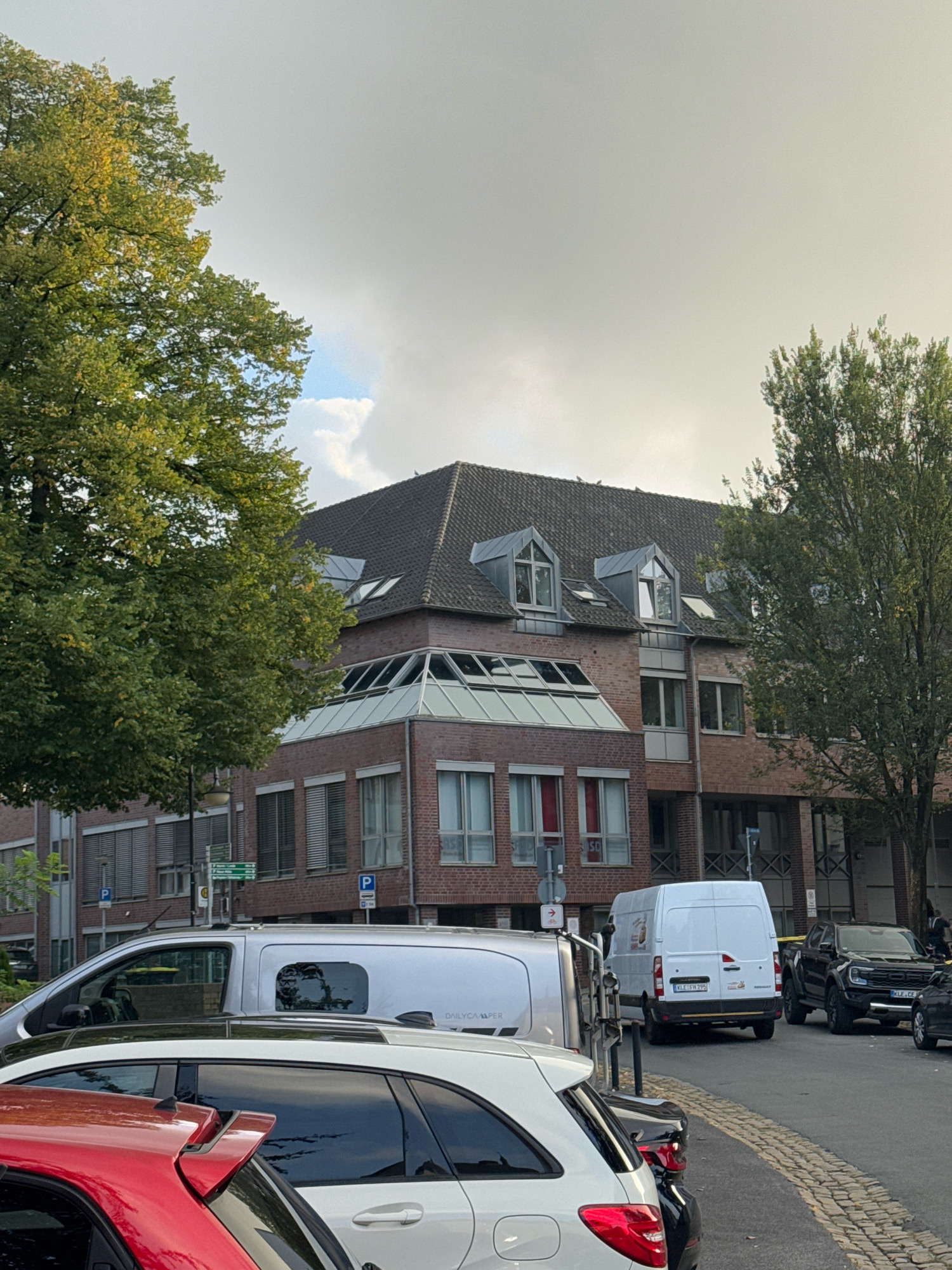 This image captures an exterior view of a brick building in the city of Cleves, Germany. The building has a prominent dark tiled roof with several dormer windows. Part of the building features a modern glass-paneled section. In front of the building, on a street paved with cobblestones, several vehicles are parked. These include a white van, a black pickup truck, and the rear ends of a white SUV and a red car. The environment appears to be an urban setting with trees present. The sky is overcast.