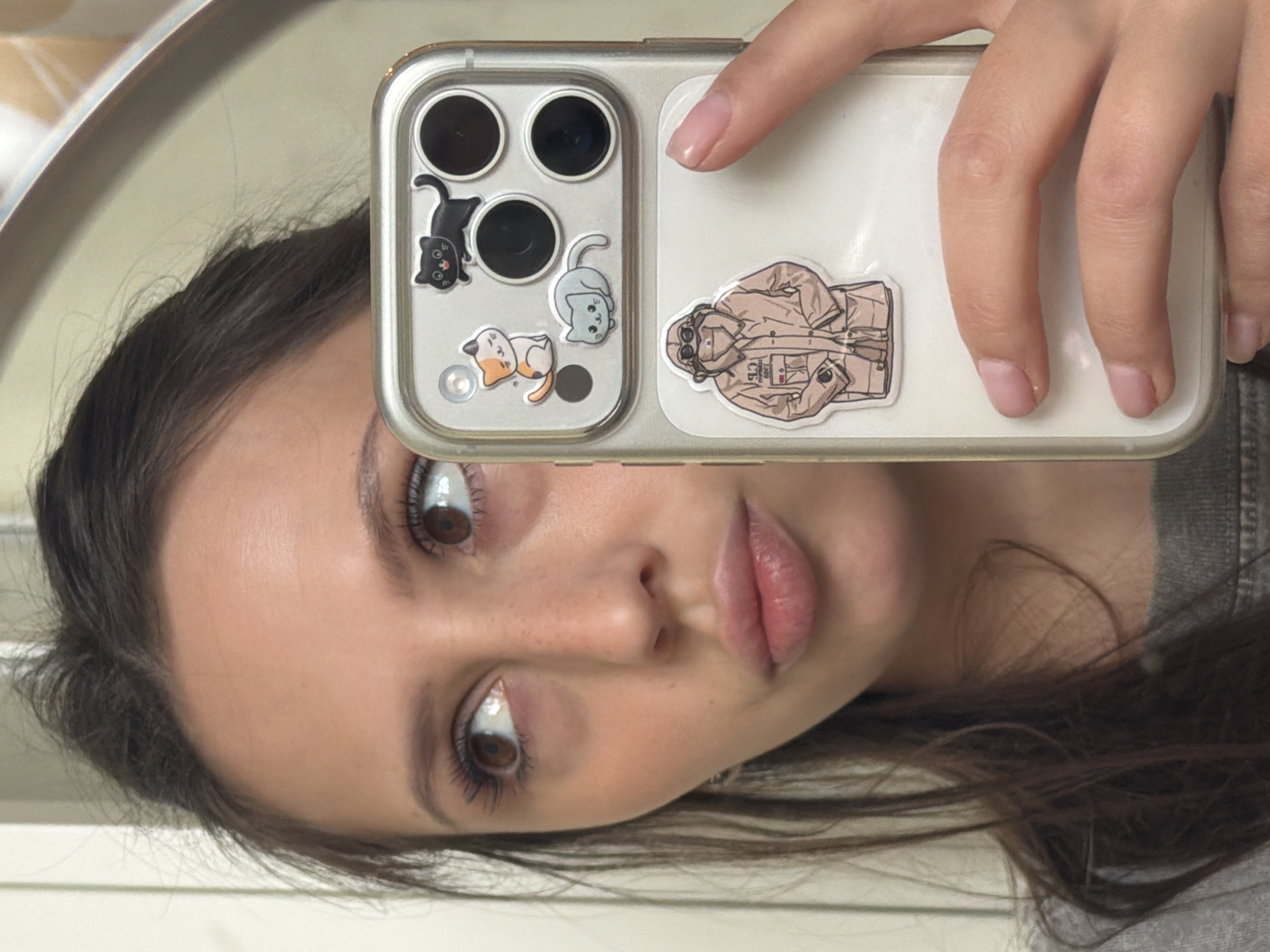 This is a vertical, close-up photograph of a young woman taking a selfie in a mirror. She is looking at the camera with her mouth slightly pursed. Her face is turned to her right, and her eyes are wide open. She has dark brown hair, which is visible around her face and falling over her shoulder. She appears to be wearing makeup, with noticeable mascara and lip gloss.
She is holding a smartphone in her right hand, the camera facing her. The phone case is clear and has several stickers on it. One sticker features a cartoonish drawing of a beige jacket with a hood and goggles. Another section of the phone case has three circular camera lenses and a flash. There are also three small cartoon cat stickers on the phone case.
The background of the image shows a mirror reflection, with a blurred silvery frame visible on the left. The lighting suggests it is daytime, likely indoors. The overall mood is casual and playful, common for a selfie.
There is no visible text in the image. The context provided indicates the location is Khimki, Russia.