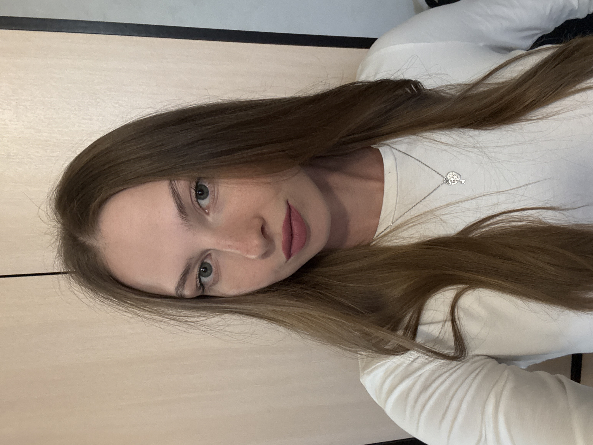 A young woman with long, brown hair is shown in a selfie. She is wearing a white, long-sleeved shirt and a silver necklace with a heart and key charm. Her eyes are light green and she has light skin with freckles. Her lips are painted a soft pink. The background appears to be an indoor setting with a wooden wall. The lighting suggests it is daytime. The image is rotated 90 degrees counter-clockwise.