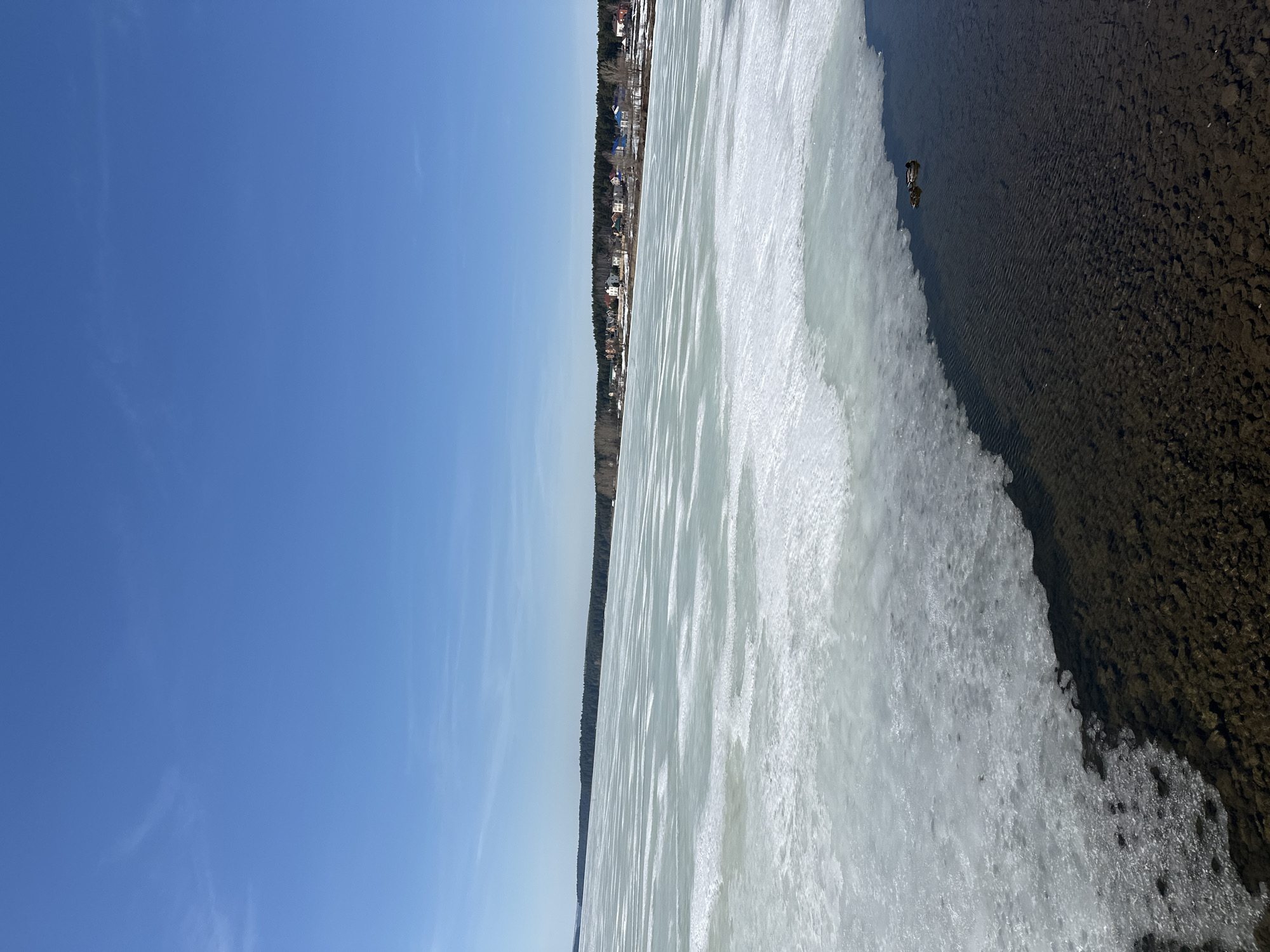The image depicts a partially frozen body of water, possibly a lake or river, with the city of Votkinsk, Russia, visible on the far shore. The left side of the image shows a clear, blue sky. A thin line of trees separates the shoreline of the body of water from the city. Patches of ice are visible with areas of open water. The right side of the image shows the shoreline, with some water. Two ducks are also visible in the water. The image is shot in the daytime with clear lighting.