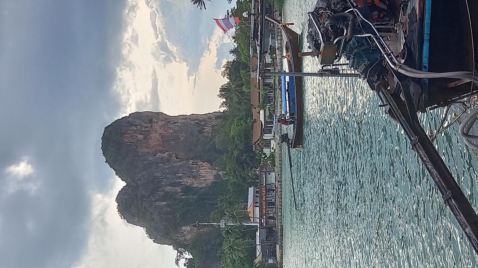 The image shows a view of the waterfront in Ban Ao Nang, Thailand. In the foreground is part of a boat, with an outboard motor and mechanical components visible. The water stretches from the boat to the town, which is built along the shoreline. Dominating the background is a large limestone karst mountain. The sky above is partly cloudy. A Thai flag is visible in front of a building. The water surface shows gentle ripples and reflections of light. A shop sign reading "BEACH OUTLETI" is visible.