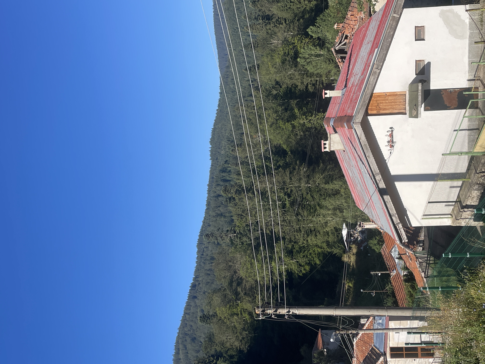 The image depicts a rural scene in Manastir, Bulgaria, characterized by a clear blue sky and a dense forest covering a hillside. In the foreground, a white house with a red roof is prominent, featuring a wooden door and two small windows. A utility pole with multiple wires stands to the left of the house, connecting to other structures in the vicinity. Several other houses with red tile roofs are visible, some appearing older or in disrepair. A green fence runs along the bottom of the image, with some vegetation and wildflowers growing near it. The overall impression is that of a quiet, mountainous village.
