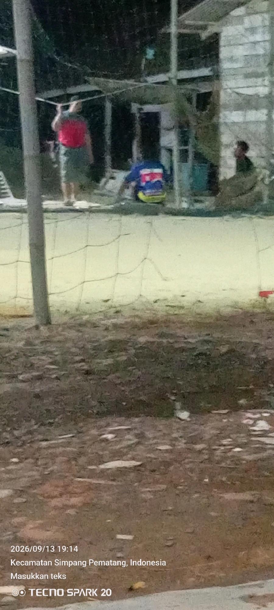 An outdoor scene at dusk shows several people near a building. In the foreground, a mesh fence stretches across the lower portion of the frame, with a gravelly, uneven ground surface below. In the midground, a person wearing a red shirt and green pants stands near a pole on the left. To the right of this person, another individual is seated on the ground, wearing a blue, white, and red striped shirt and yellow pants. Further to the right, a third person is partially visible, wearing a dark shirt and looking to the right. The background reveals a building with a corrugated roof and light-colored walls. The overall lighting suggests late evening or twilight. The image is dated 2026/09/13 at 19:14 and is located in Simpang Pematang, Indonesia, with "Masukkan teks" text also present. The bottom right corner displays "TECNO SPARK 20".
