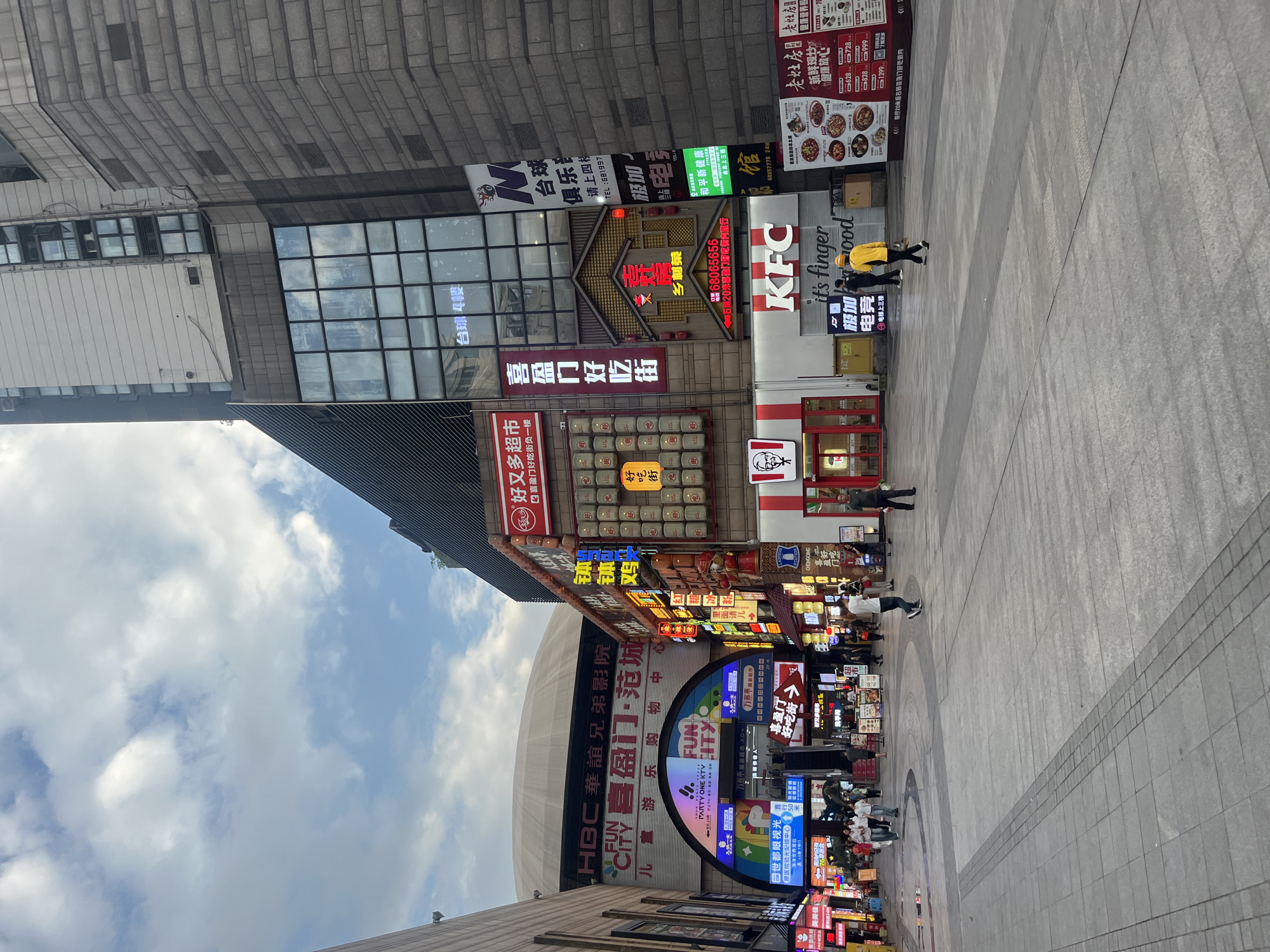 The image captures a vibrant commercial street or plaza in Jiulongpo District, China, under a bright, cloudy sky. The scene is bustling with numerous multi-story buildings featuring a dense array of colorful and brightly lit signs, predominantly in Chinese characters.
Several people are visible, some walking across the wide, grey-tiled pavement, while others are near the storefronts. A prominent KFC restaurant with its iconic red and white logo and "it's finger lickin' good" slogan stands out.
Notable establishments and services indicated by visible text include "HBC华谊兄弟影院" (HBC Huayi Brothers Cinema), "喜盈门好吃街" (Xiyingmen Delicious Street), "又好又多超市" (Youhao Youduo Supermarket), "FUN CITY", "PARTY ONE KTV", "极加电竞" (Jijia E-sports), and "台球" (Billiards). Other signs advertise "和平新健康" (Heping New Health), "老灶房" (Laozaofang) with a menu displaying prices, "乡村茶" (Country Tea), and "世都眼视光" (Shidou Optometry). Directions such as "直行50米" (Go straight 50 meters), "电梯上三楼" (Elevator to 3rd floor), and mentions of "负一楼" (Basement 1st floor) are also present. The architecture is modern, with large windows and textured grey building facades. The overall impression is that of a lively urban shopping and entertainment district during the daytime.