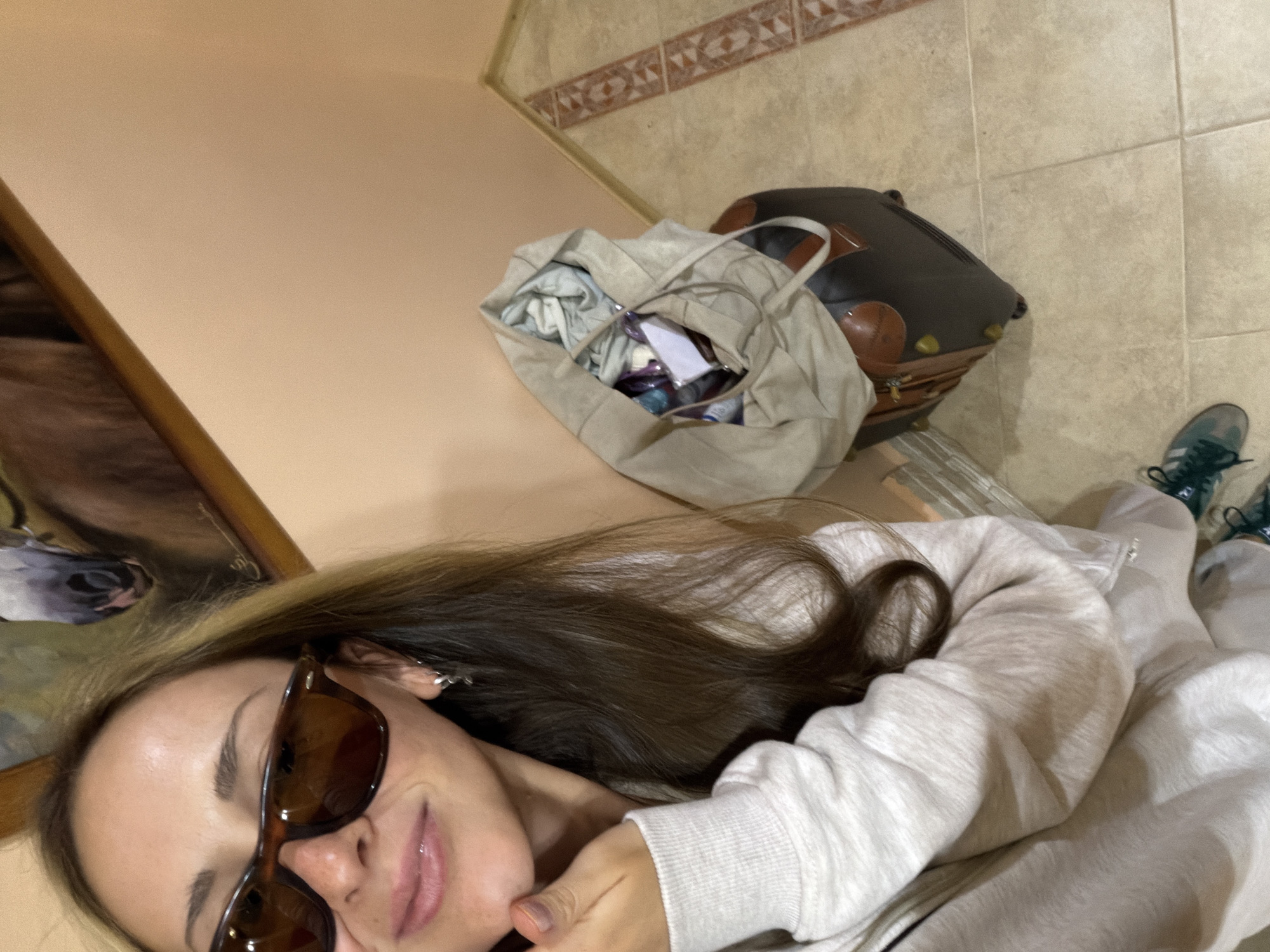 The image is a high-angle shot of a woman in sunglasses, wearing a light-colored sweatshirt, with her hair partially obscuring her face. To the woman's left is a painting, and to her right are a large beige tote bag and a brown leather suitcase. In the background, the flooring appears to be light-colored tile, and there is decorative tile along the wall. The scene appears to be indoors, potentially in a home or hotel room. There is no indication of the city Kamyshin, Russia.