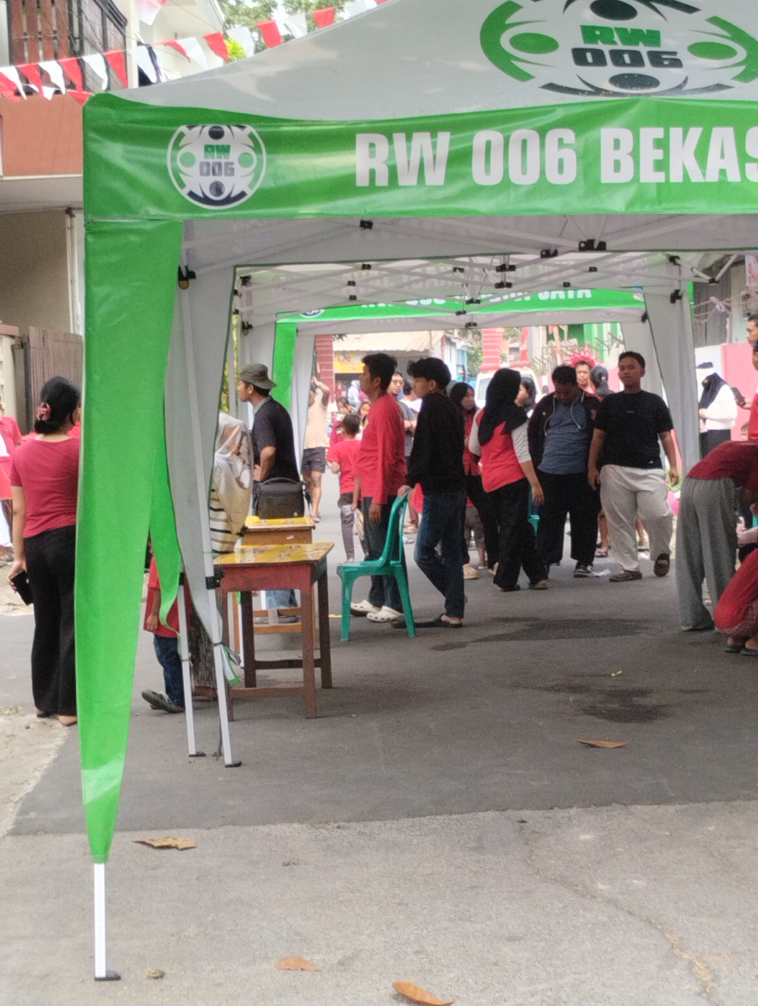 In Bekasi, Indonesia, a group of people are gathered under a green and white tent that reads "RW 006 BEKASI". A woman in a red shirt and black pants stands on the left, facing away from the camera. To her right, a man in a hat and dark shirt is speaking with another person. In the background, more individuals are seen milling around, some wearing red shirts. The ground is paved with asphalt and concrete, and the overall scene suggests a community event or gathering.