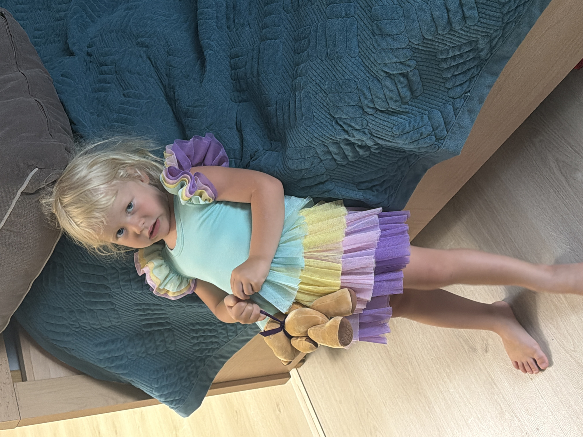 A young Caucasian girl with blonde hair and blue eyes stands barefoot on a light wood floor. She is wearing a light blue leotard with multi-colored ruffles on the shoulders and a layered tulle skirt in pastel shades of yellow, lavender, and purple. She is holding a small, light brown teddy bear by its ribbon in her hands. The girl is positioned next to a bed with a textured, dark teal quilted comforter and a beige headboard. The setting appears to be indoors, possibly a bedroom, with natural light illuminating the scene from the left. The image does not contain any specific identifiable landmarks or clues that would place it in Sochi, Russia.