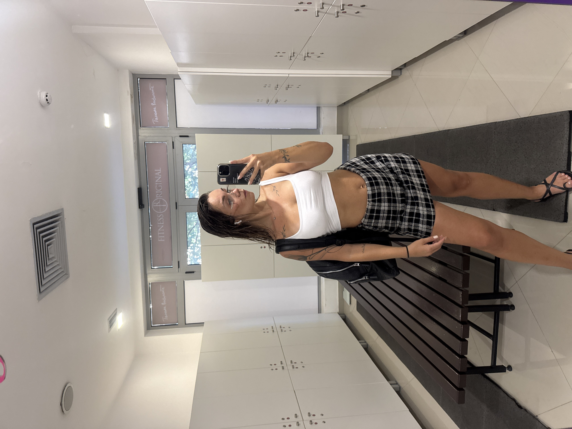 A woman takes a selfie in a gym locker room. She is wearing a white crop top and a black and white plaid skirt. She has tattoos on her arms and chest. She is holding a smartphone in her right hand and looking at the camera. To her left, there is a wooden bench with a black bag on it. In the background, there are white lockers and a window with a view of trees. The floor is tiled with a gray mat in front of the bench. The lighting suggests it is daytime. The text "FITNESS ORIGINAL" is visible on a window in the background. The location is Budva, Montenegro.