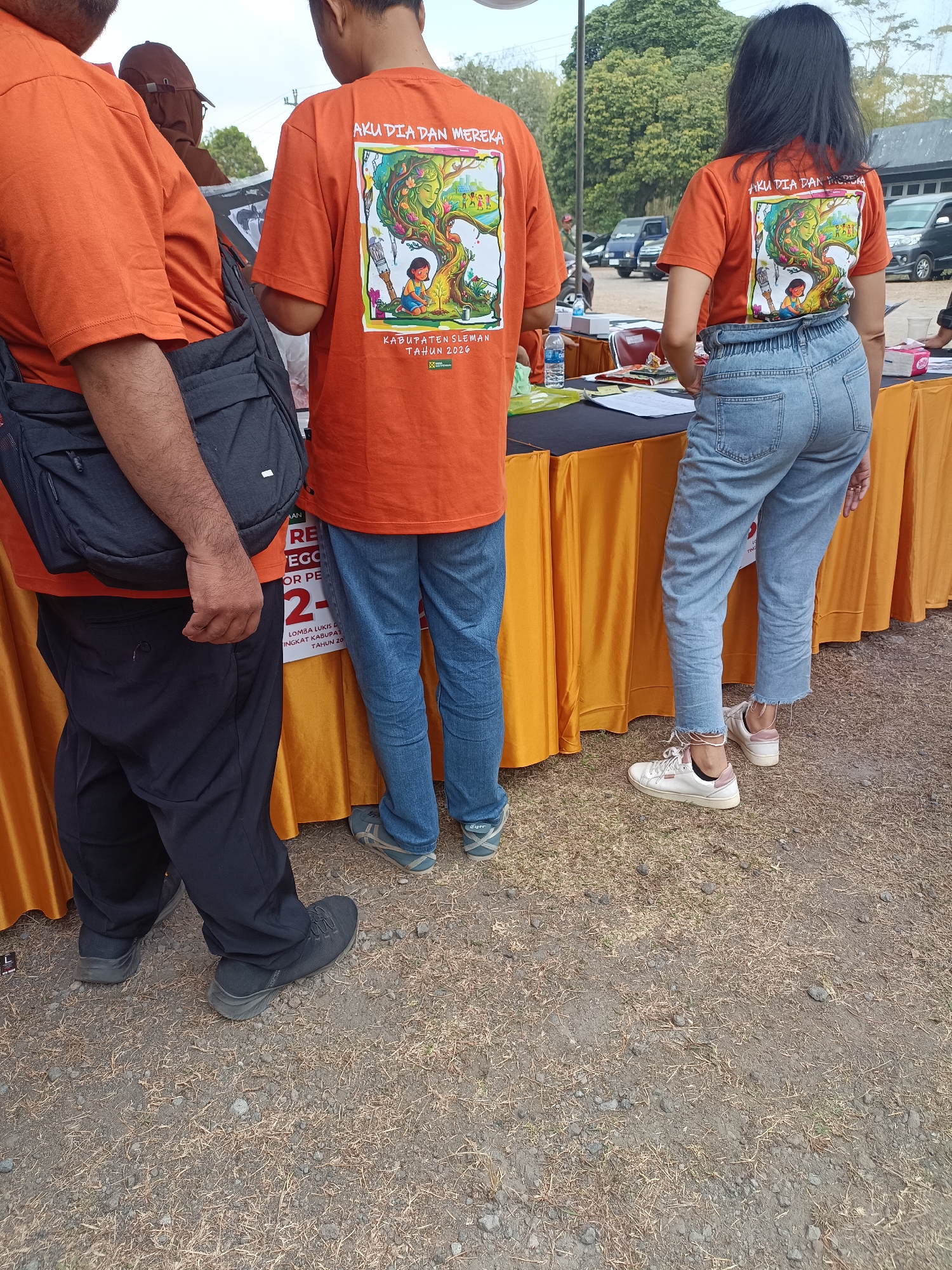 Three individuals, two men and one woman, are standing behind an orange draped table. The man on the left is wearing an orange t-shirt and black pants. He has a dark bag slung over his shoulder. The man in the middle is wearing an orange t-shirt with a graphic design on the back. He has blue jeans on. The woman on the right is also wearing an orange t-shirt with a similar graphic design on the back. She is wearing light blue jeans. On the table are various items including books, bottles of water and papers. The background shows trees, cars and a building, suggesting an outdoor event. The scene appears to be in Hargobinangun, Indonesia based on the visible text on the t-shirts which includes "KABUPATEN SLEMAN TAHUN 2020".