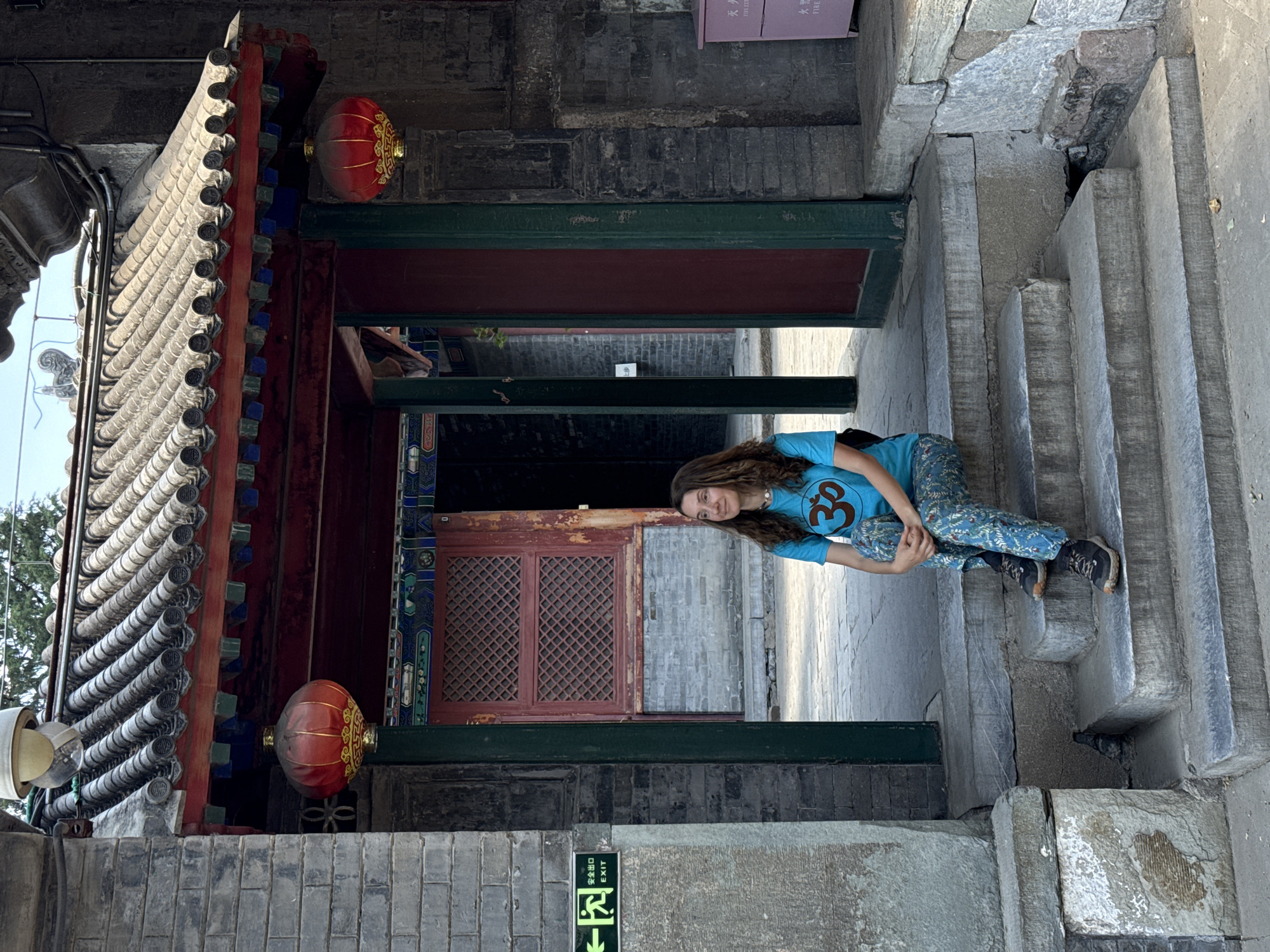 A person is sitting on stone steps inside an architectural structure with red lanterns and ornate details. The location is the Xicheng District of China. The person is wearing a blue shirt with a symbol on it and patterned pants. An exit sign is visible. The surrounding architecture is primarily stone and brick.