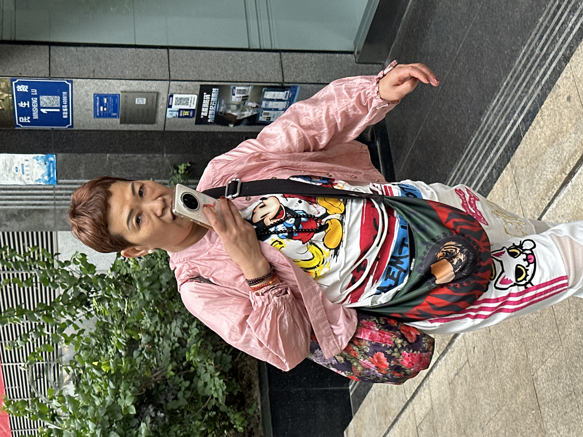 A woman is seen holding a cell phone in her left hand while speaking, positioned outdoors. She is wearing a pink blouse over a T-shirt decorated with cartoon characters and a black strap over her shoulder; she is also carrying a floral handbag. In the background, there is greenery and the side of a building. A blue street sign is visible, labeled "MINSHENG LU", indicating that the location is in the Yuzhong District of Chongqing, China.