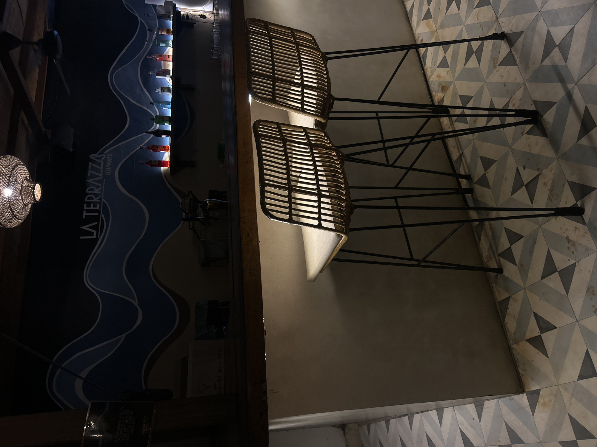 The image depicts the interior of a bar or restaurant with a focus on the bar area and seating. Two rattan bar stools with metal legs are positioned in front of a textured bar counter. The counter is lit from below, casting a warm glow. Behind the counter, a shelf displays an assortment of bottles, likely spirits, illuminated by a soft light. A dark blue wall with a wavy blue and white mural serves as the backdrop, featuring the text "LA TERRAZZA ULUWATU". A ceiling fan and a woven light fixture are visible above. The floor is tiled with a geometric pattern of light and dark grey, blue, and beige triangles. The overall ambiance suggests a dimly lit, stylish establishment. The location is identified as Kuta Selatan, Indonesia.