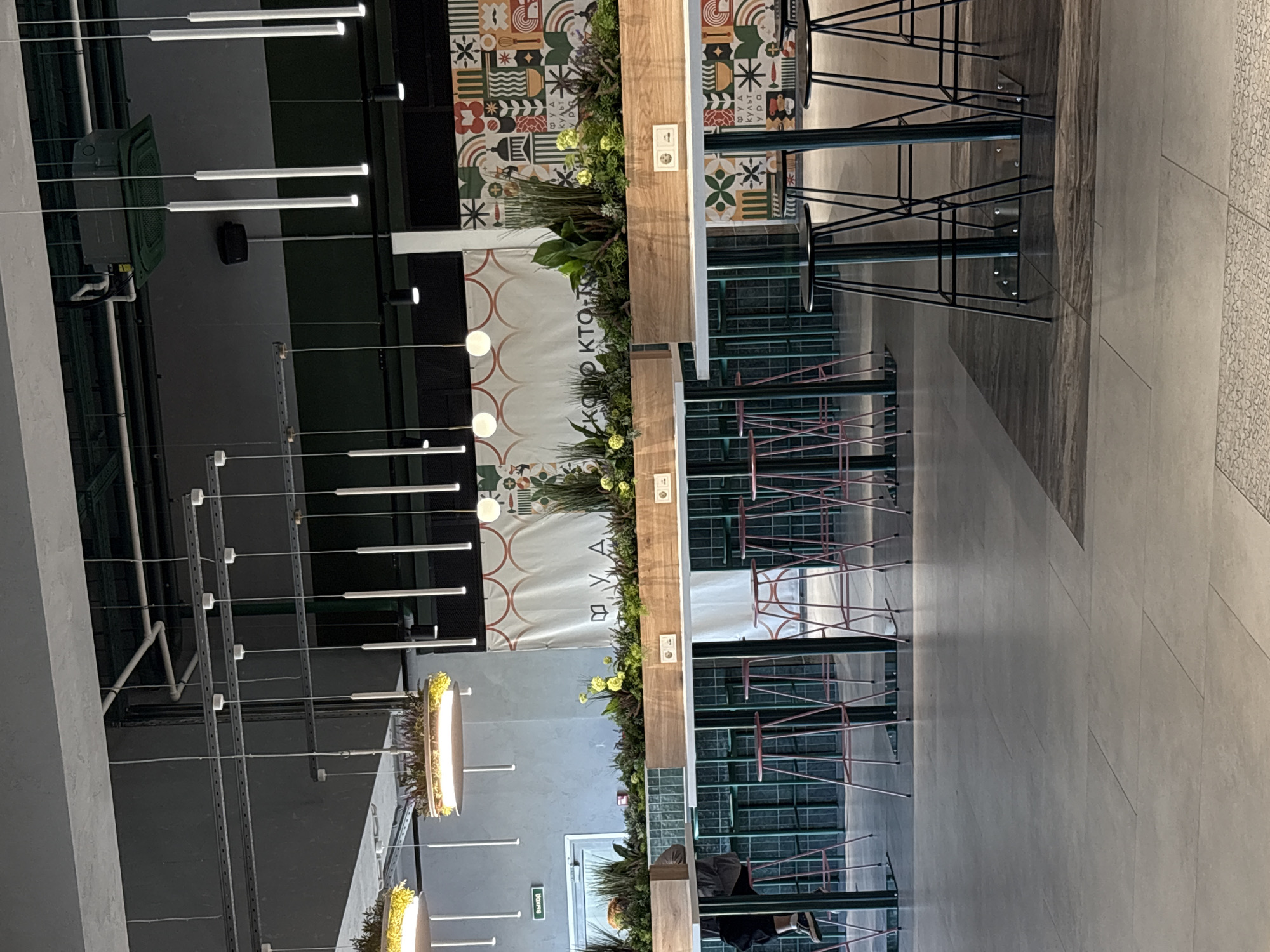 The image depicts the interior of a modern cafe or restaurant located in Saint Petersburg, Russia. In the foreground, high tables with black stools are arranged. Behind these, a long counter area is visible, featuring a wooden planter filled with lush green plants and yellow flowers, with white electrical outlets integrated into the counter. The counter is backed by a wall with a dark green tiled pattern. In the background, a large, colorful mural with a geometric and patterned design adorns the wall, interspersed with Cyrillic text. Multiple modern pendant lights hang from the ceiling. To the left, a door with a green "EXIT" sign is visible, suggesting this might be an establishment within a larger building or complex. The overall ambiance is contemporary and vibrant, with an emphasis on natural elements and modern design.