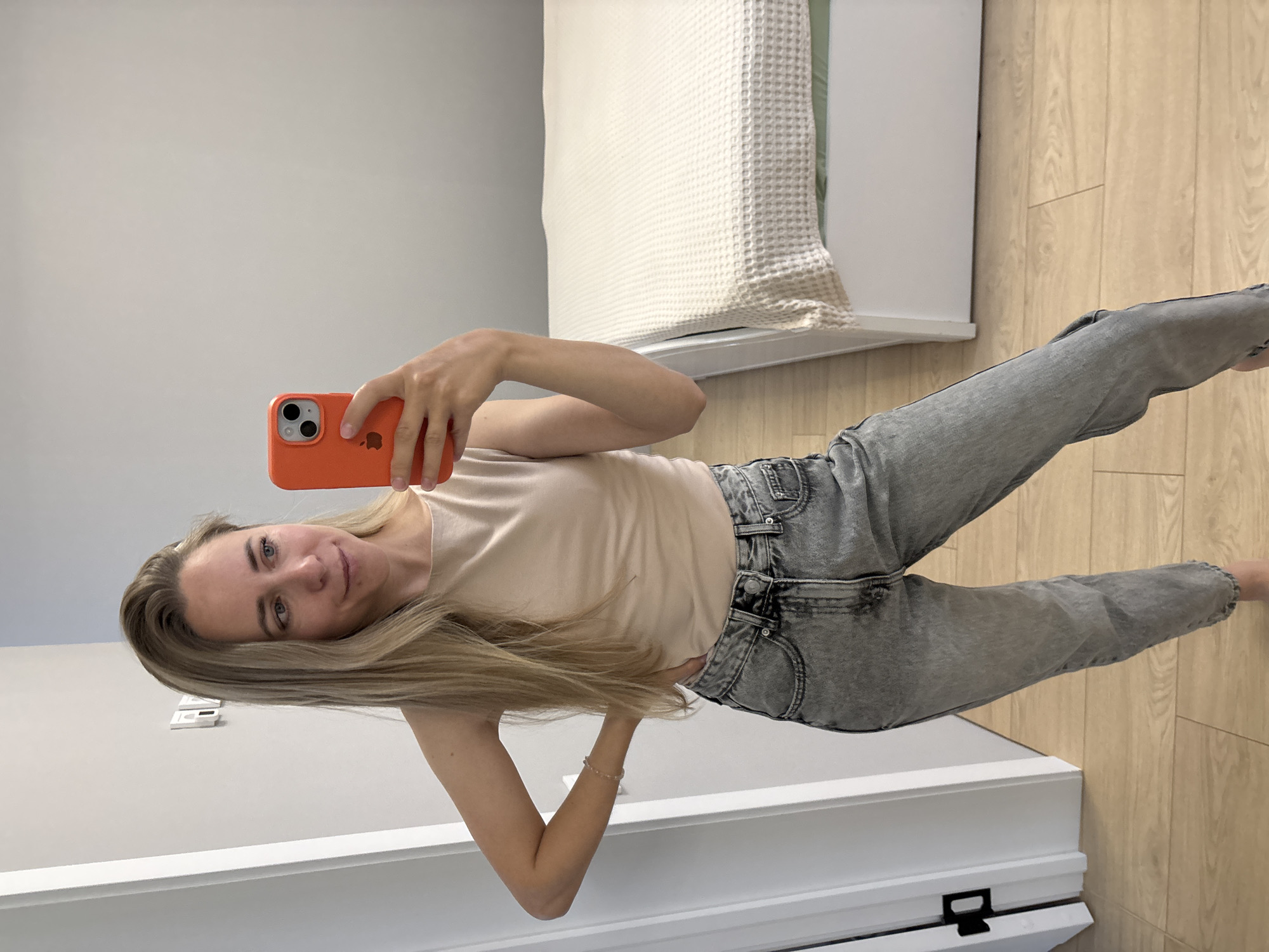 A young woman with long blonde hair is taking a mirror selfie in what appears to be a bedroom. She is wearing a light beige sleeveless top and grey, high-waisted, loose-fitting jeans. She is holding an orange iPhone in her right hand, which is angled to capture her reflection. Her left hand is on her hip. In the background, a white bed with a light-colored textured blanket is visible, along with a white door frame to the left. The flooring is light wood laminate. There is no indication of the specific location within the city of Chaikovsky, Russia.
