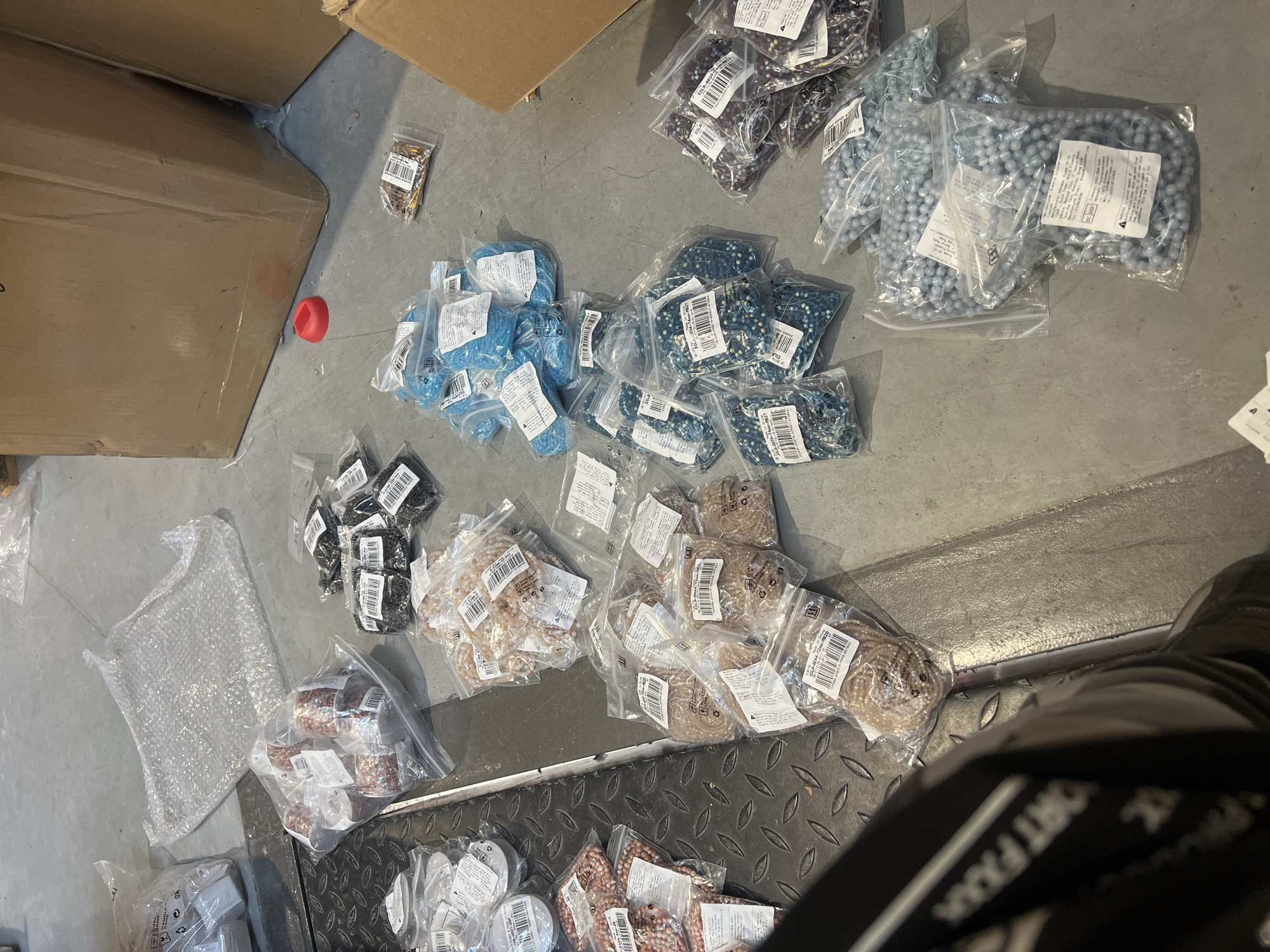 The image depicts a variety of packaged beads of different colors and types scattered on the floor, alongside cardboard boxes and packing materials. The beads are in plastic bags, and some have labels attached. The floor surface appears to be concrete in some areas and metal with a tread plate pattern in others. A portion of a person's leg is visible in the lower right corner. There is also a red plastic object near one of the boxes. It is impossible to determine that the location is in the city of Burgas, Bulgaria with the given images.
