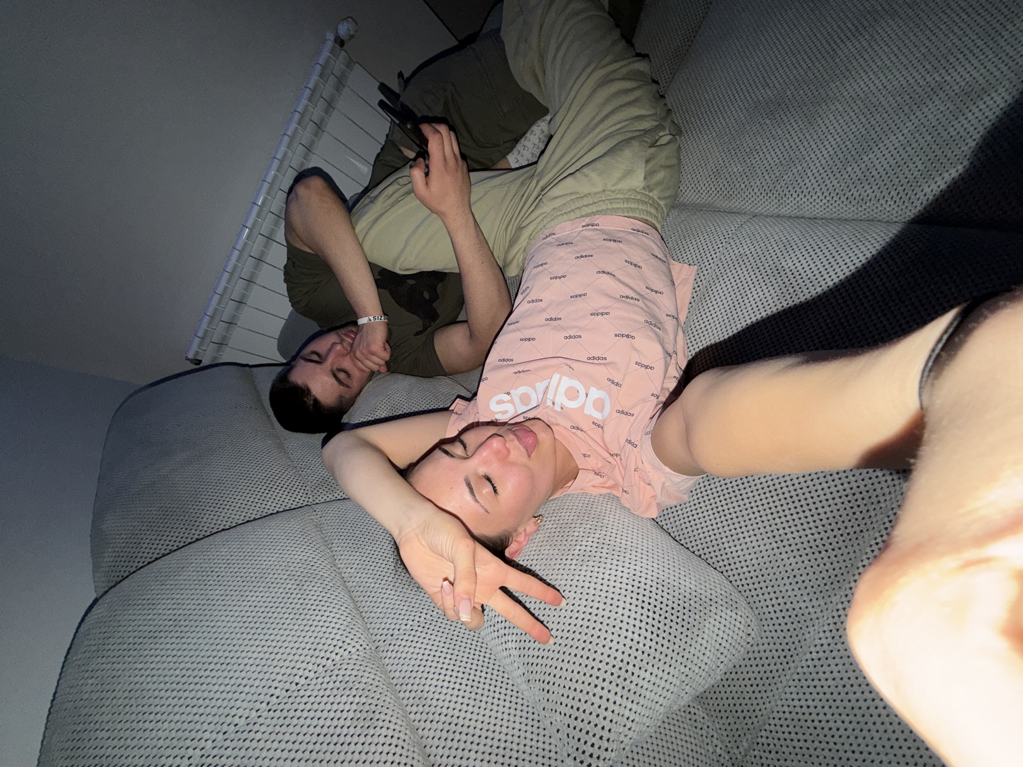 A photograph shows two people reclining on a textured gray couch. One individual, presumably female, is closer to the camera, lying on their back. They are wearing a pink t-shirt with the Adidas logo pattern and are making a peace sign with one hand while sticking their tongue out. Their head is turned slightly to the left. Part of an arm and hand is very close to the camera, appearing blurred.
The second person, presumed male, is lying behind the first, also on their back. He is wearing a dark green t-shirt and beige pants. He is using a phone, held in his hands. A white radiator is visible in the background.
The location is presumed to be indoors, possibly in a home in Buhovo, Bulgaria, given the user's location. The lighting seems to be dim, with some shadow play on the couch. The setting suggests a casual, relaxed environment.