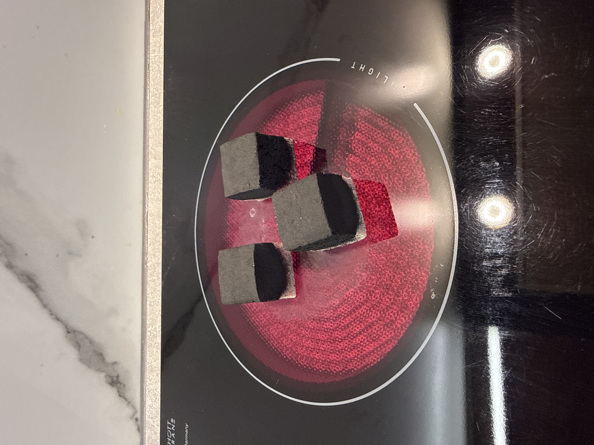 The image shows three black, cubical hookah coals resting on a red, circular electric stovetop burner. The burner is illuminated and appears to be hot. The stovetop is black glass with white circular markings indicating the burners. In the upper left corner, a portion of a marble countertop is visible. The overall scene is a close-up shot of the stovetop and coals, with no indication of a specific city or location.