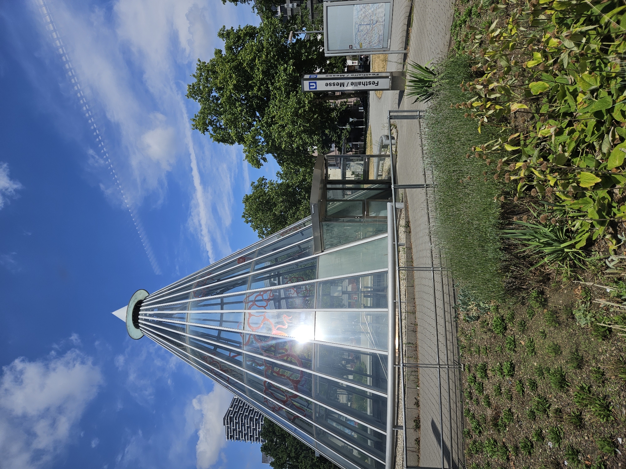The image depicts the entrance to the U-Bahn station Festhalle/Messe in Frankfurt, Germany. The main subject is a striking glass pyramid structure, a modern architectural design with a conical shape, serving as the station's entrance. The glass panels of the pyramid are partially obscured by red artistic elements, possibly graffiti or etched designs, and the bright sunlight reflects off the glass.
Inside the pyramid, stairs are visible leading down, indicating access to the underground platform. A small, glass-enclosed structure, possibly a shelter or ticket booth, is situated next to the pyramid. A tall, blue and white sign with the "U" logo for public transport and the station name "Festhalle / Messe" stands prominently near the entrance. To the right of the sign, a public information board displaying a map is mounted.
The surrounding environment includes lush green trees, a blue sky with scattered white clouds, and a contrail from an aircraft. The ground is paved with interlocking bricks in some areas and covered with gravel and vegetation in others. A metal railing separates the paved walkway from a planted area in the foreground, which features various types of plants, including lavender and other leafy green shrubs. In the background, a modern, high-rise building with a distinctive facade is visible. There are no people present in the image. The scene is a bright, sunny day.