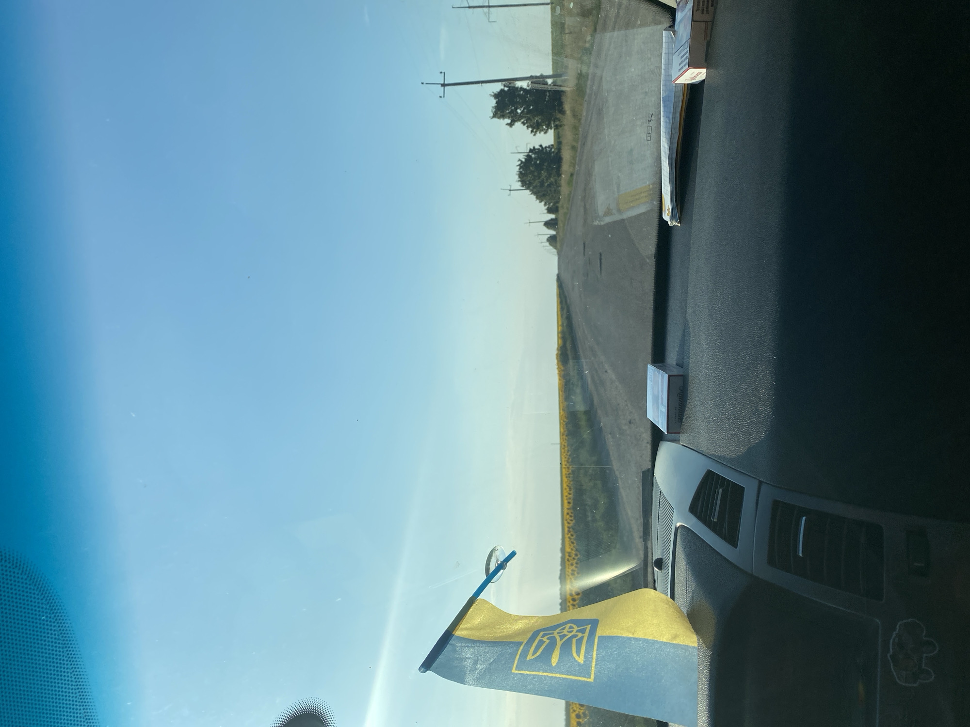 The image depicts a view from inside a vehicle looking out through the windshield. In the foreground, on the dashboard, is a small Ukrainian flag attached to the windshield with a suction cup. To the right of the flag are two packs of cigarettes. The road ahead stretches into the distance, lined by telephone poles and trees. On either side of the road are fields, with a large field of sunflowers visible to the left. The sky is clear and blue. The location is stated to be in the city of Maiske, Ukraine.