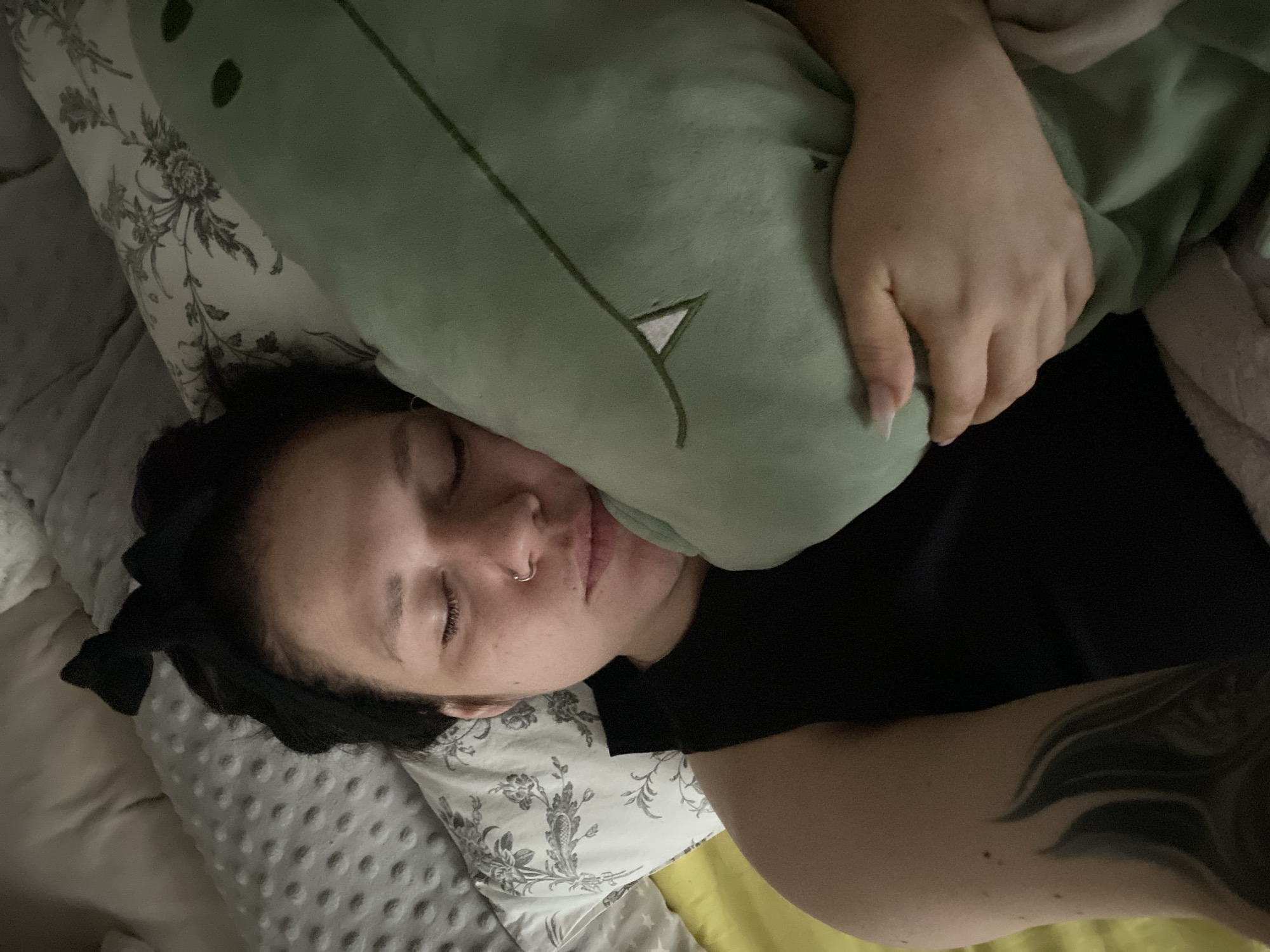A person is sleeping in bed, clutching a green plush dinosaur pillow. The person is lying on their side with their eyes closed. They have a nose piercing and dark hair pulled back with a bow. They are wearing a black shirt. Part of their arm and a visible tattoo is to the right of the image. The bed has a floral patterned pillowcase. The sheets are varying textures and colors. The photo was taken indoors. The location is not discernible based on the information in the image and it can not be determined whether this photo was taken in the city of Chaikovsky, Russia.