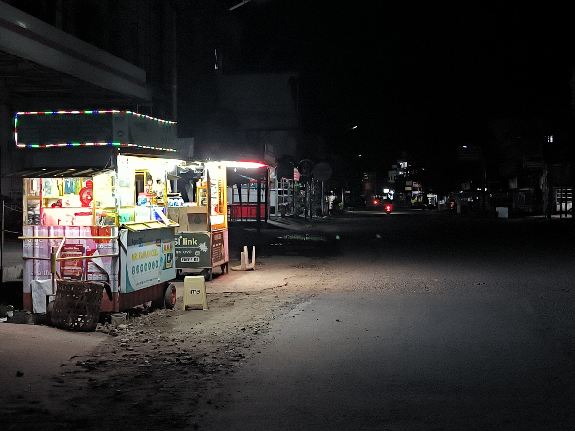 This nighttime image depicts a street scene in Lhokseumawe, Indonesia, dominated by two brightly lit mobile vendor carts. The cart on the left has a colorful LED strip running along its top edge. Various goods are visible on the cart, including what appears to be stacks of boxed goods. It also features a sign reading "MR. RAIHAN CELL". The second cart, positioned to the right, is smaller and also illuminated. The road is asphalt, and the area surrounding the carts is uneven with dirt and debris. In the background, streetlights illuminate a stretch of the road, revealing other buildings and structures in the distance. A vehicle with red tail lights is present further down the street. The overall scene suggests a quiet night in a city area with limited activity.