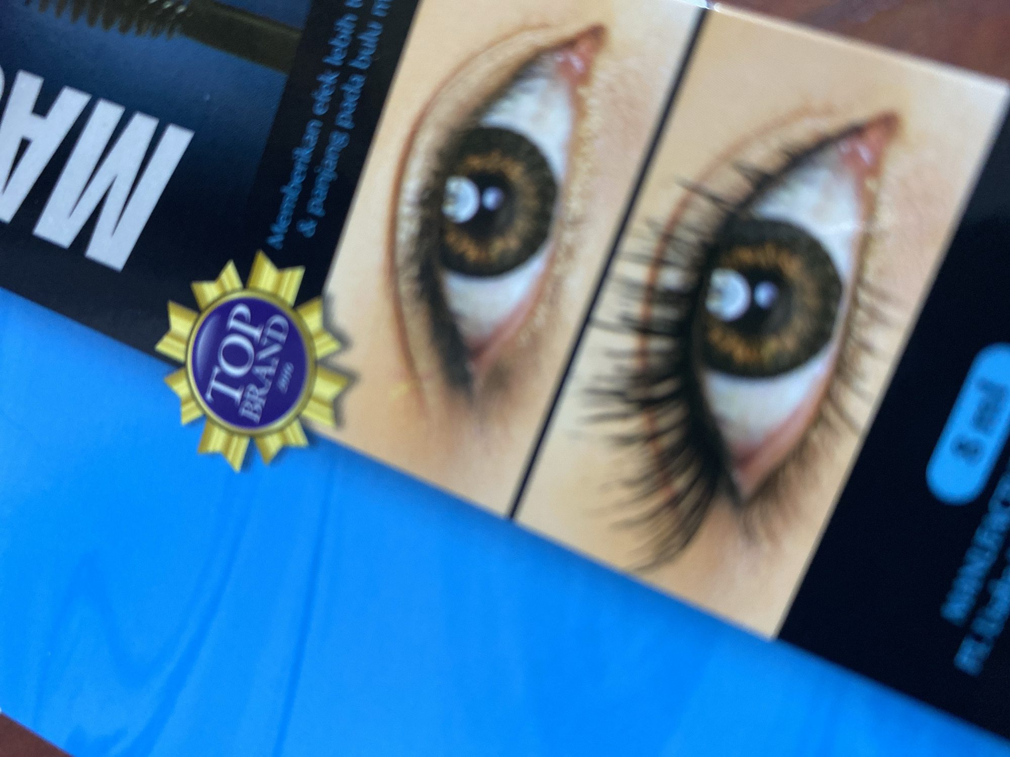 The image is a close-up, angled shot of a product package, likely mascara, displayed against a bright blue background. The package features images of a woman's eye before and after applying the product, showcasing longer, fuller lashes in the "after" image. A "TOP BRAND" award seal is visible on the left side of the package. Text on the package includes "MAS" and a descriptive phrase in Indonesian, "Memberikan efek lebih tebal & panjang pada bulu mata," which translates to "Provides a thicker & longer effect on eyelashes." A small oval shape with "8 ml" is also visible. The lighting is somewhat harsh, creating reflections on the product packaging. The blue background appears to be a textured surface. There is no discernible information about a specific location in Ta', Indonesia.