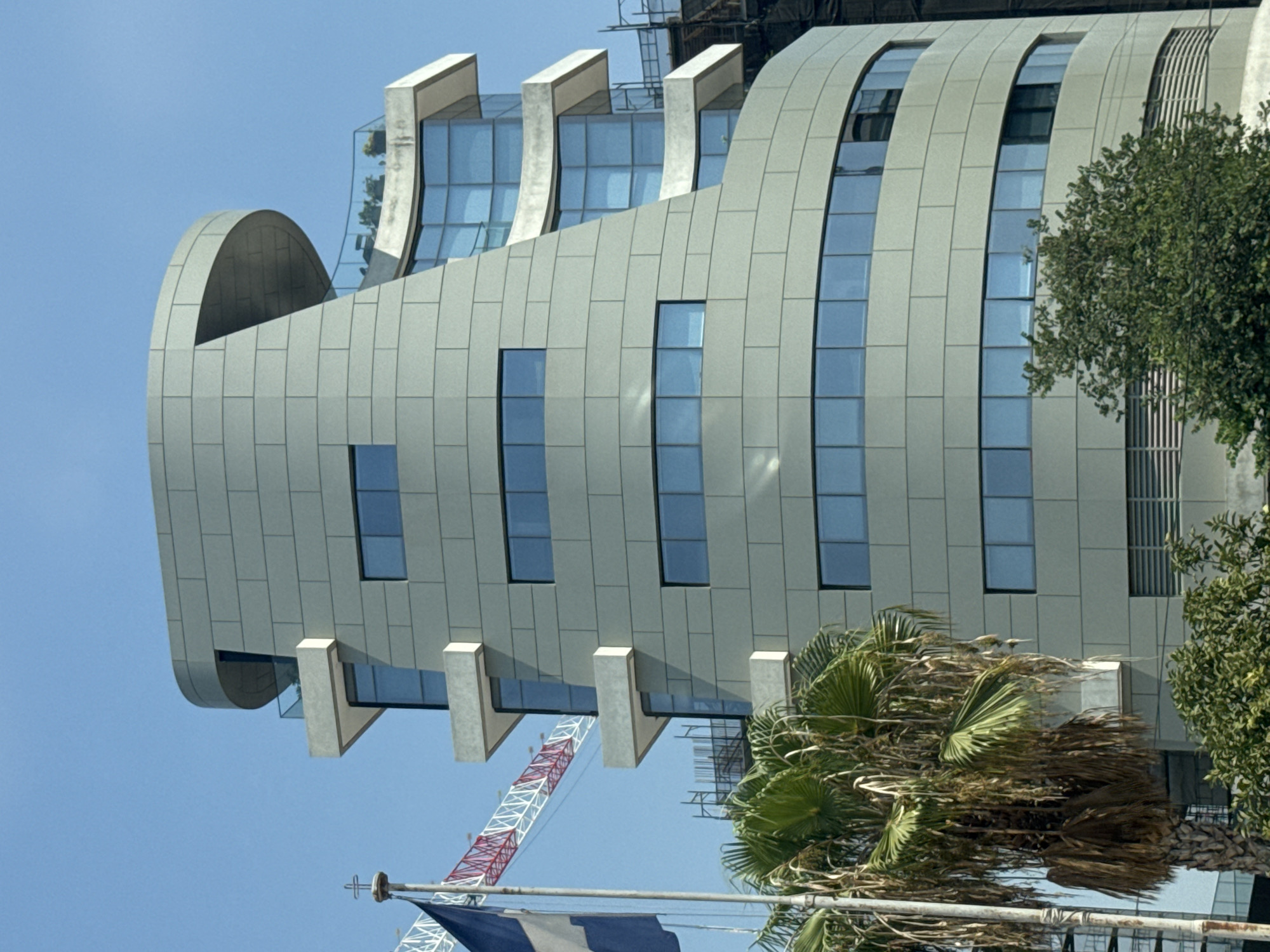 The image shows the modern architectural design of a building in Limassol, Cyprus. The building is characterized by its curved facade clad in light grey panels and numerous rectangular windows. Several cantilevered balconies jut out from the structure, some featuring glass railings and outdoor seating. To the left of the main building, another section of the structure with similar architectural elements, including balconies and glass windows, is visible. In the foreground, a palm tree with lush green fronds and some dry, hanging foliage is present. A flagpole with the flag of Greece is also visible, as is a construction crane in the background, suggesting ongoing development in the area. The scene is set against a clear blue sky.